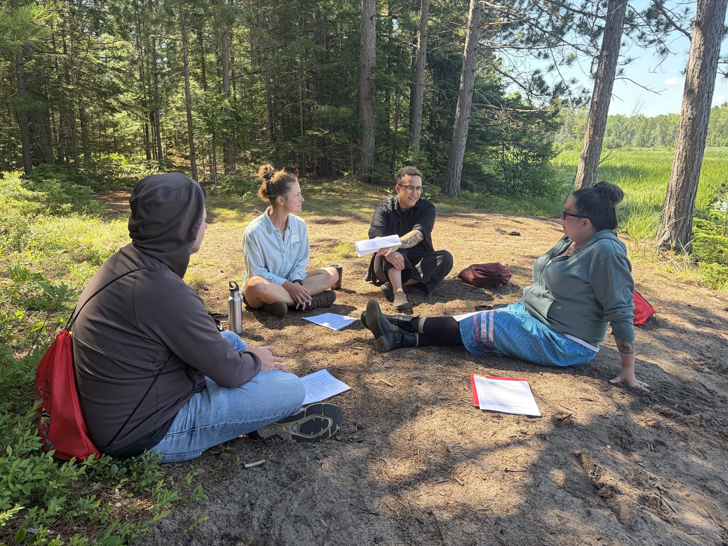Four people sitting in a circle outdoors in a forest clearing, engaged in conversation, with papers and a water bottle nearby.