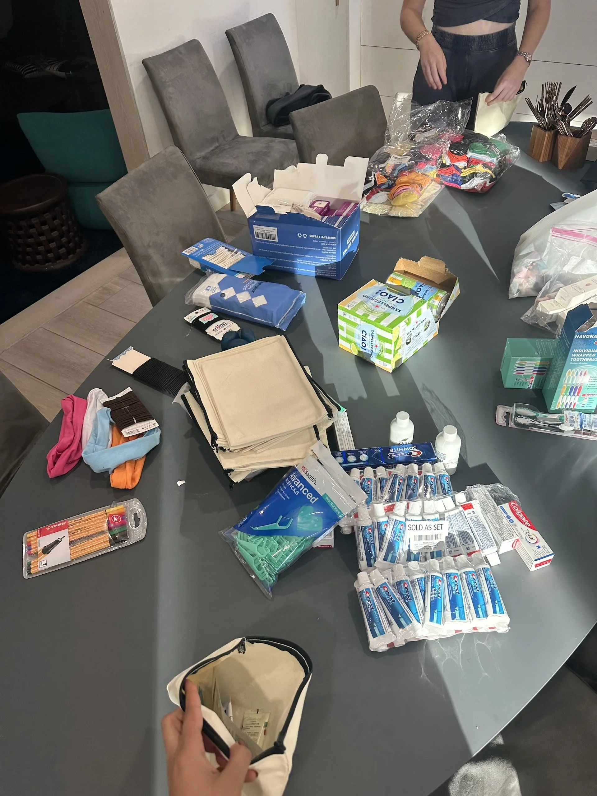 A table covered with donated supplies for LoveYOU Project care packages, including hygiene products like soap, toothpaste, and wet wipes, school supplies such as pencils and notebooks, colorful socks, canvas drawstring bags, and personal care items. 