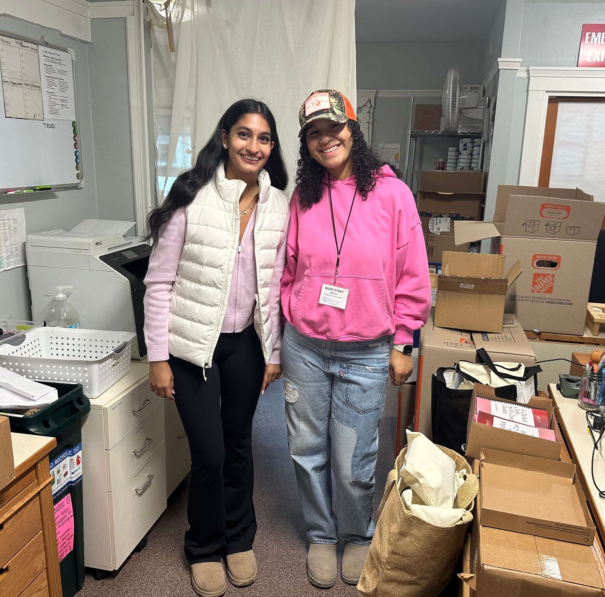Zoya from LoveYou Project and a Volunteer are standing together in a workspace with boxes of self-care items and tote bags, organizing supplies for the LoveYOU Project’s initiative to support needy women.
