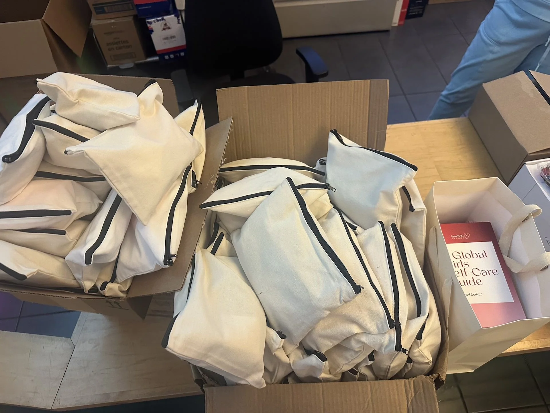 A cardboard box filled with completed LoveYOU Project care packages ready for distribution. Canvas drawstring bags are neatly packed alongside copies of the "Global Menstrual Self-Care Guide" and other resources, representing the comprehensive suppor