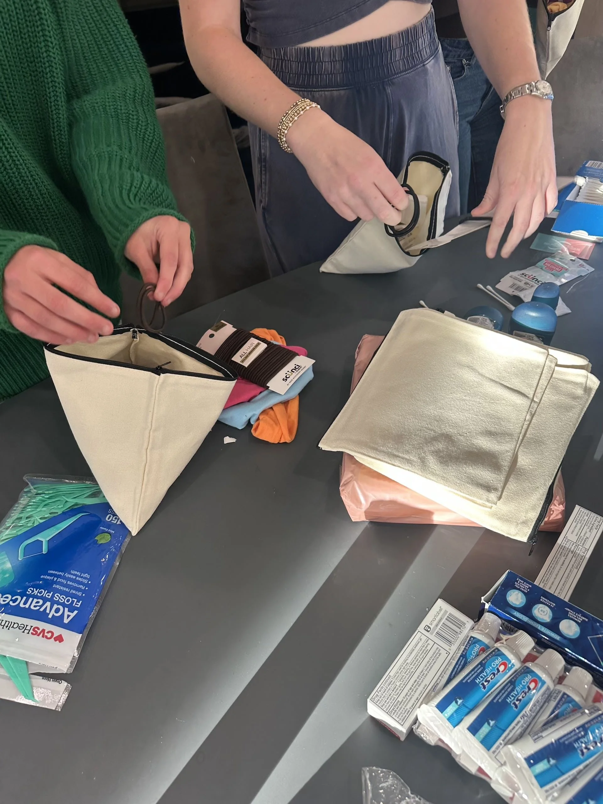 Two volunteers working side-by-side to fill canvas drawstring bags with donated items. The table displays organized supplies including colorful socks, hygiene products, and school materials. Each volunteer holds a bag as they carefully select items t