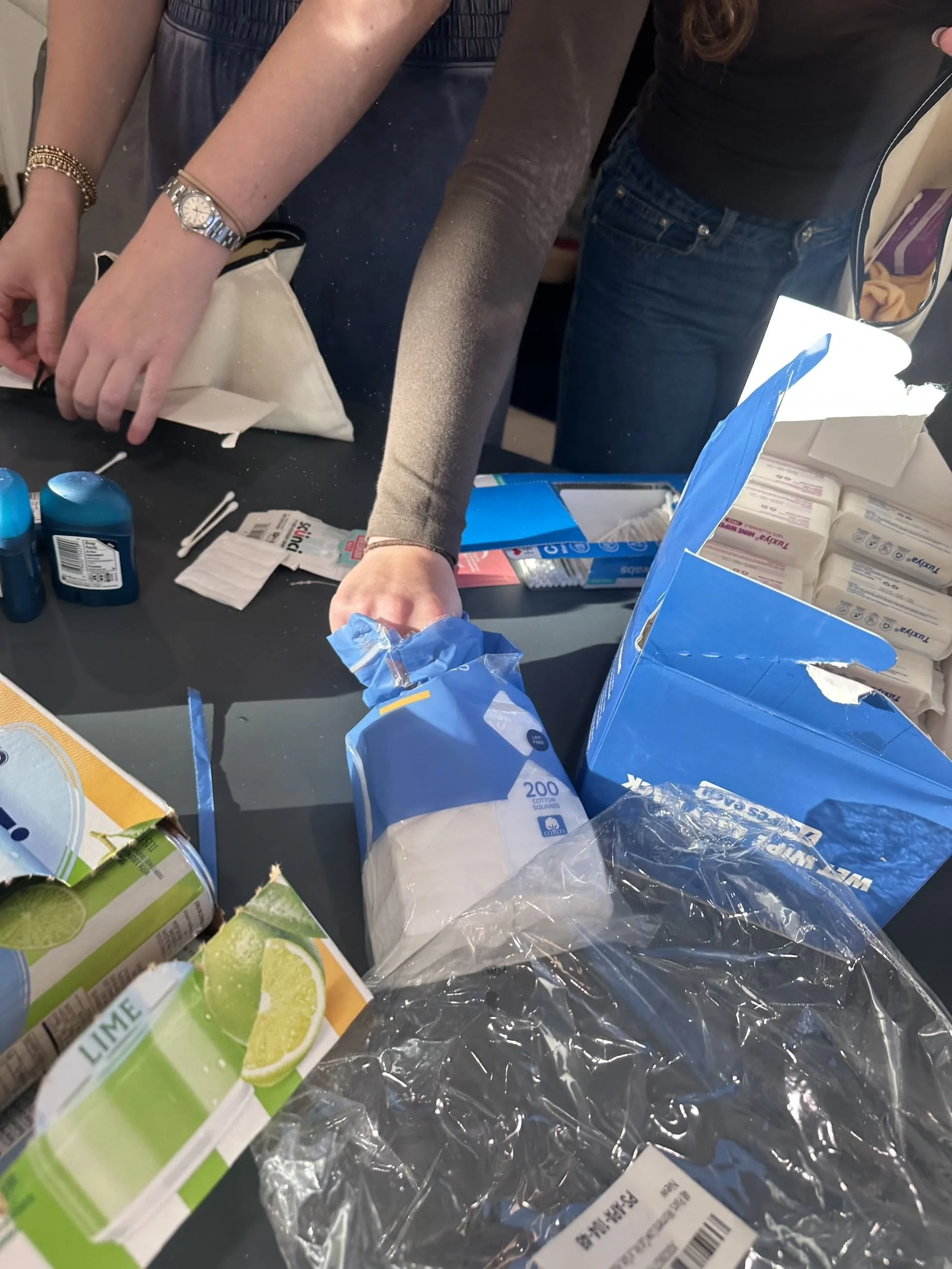 Two volunteers working side-by-side to fill canvas drawstring bags with donated items. The table displays organized supplies including colorful socks, hygiene products, and school materials. Each volunteer holds a bag as they carefully select items t