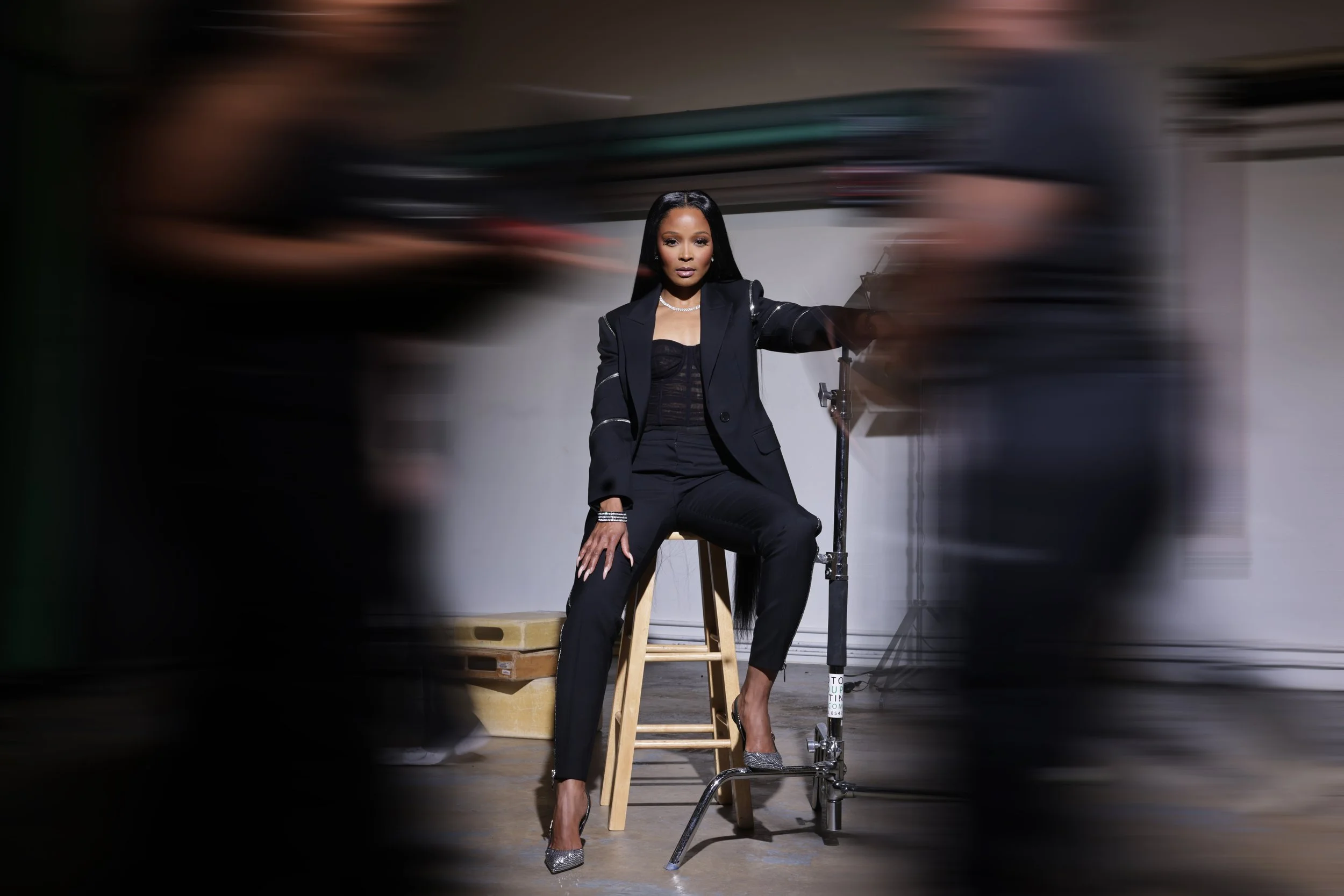 A woman dressed in a black blazer, top, and pants, sitting on a stool in a studio with a plain white and gray background. She wears heels and jewelry, and is being photographed with motion blur around her.