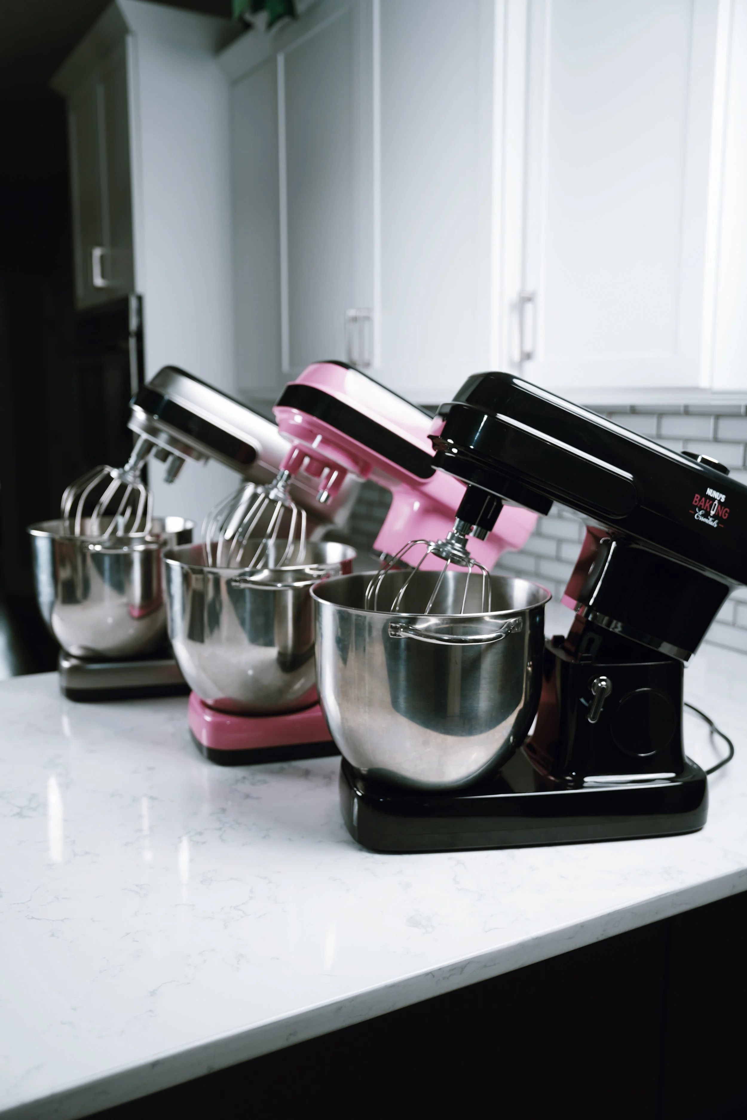 Three stand mixers in black and pink colors, each with a metal mixing bowl and beaters, placed on a white kitchen countertop.