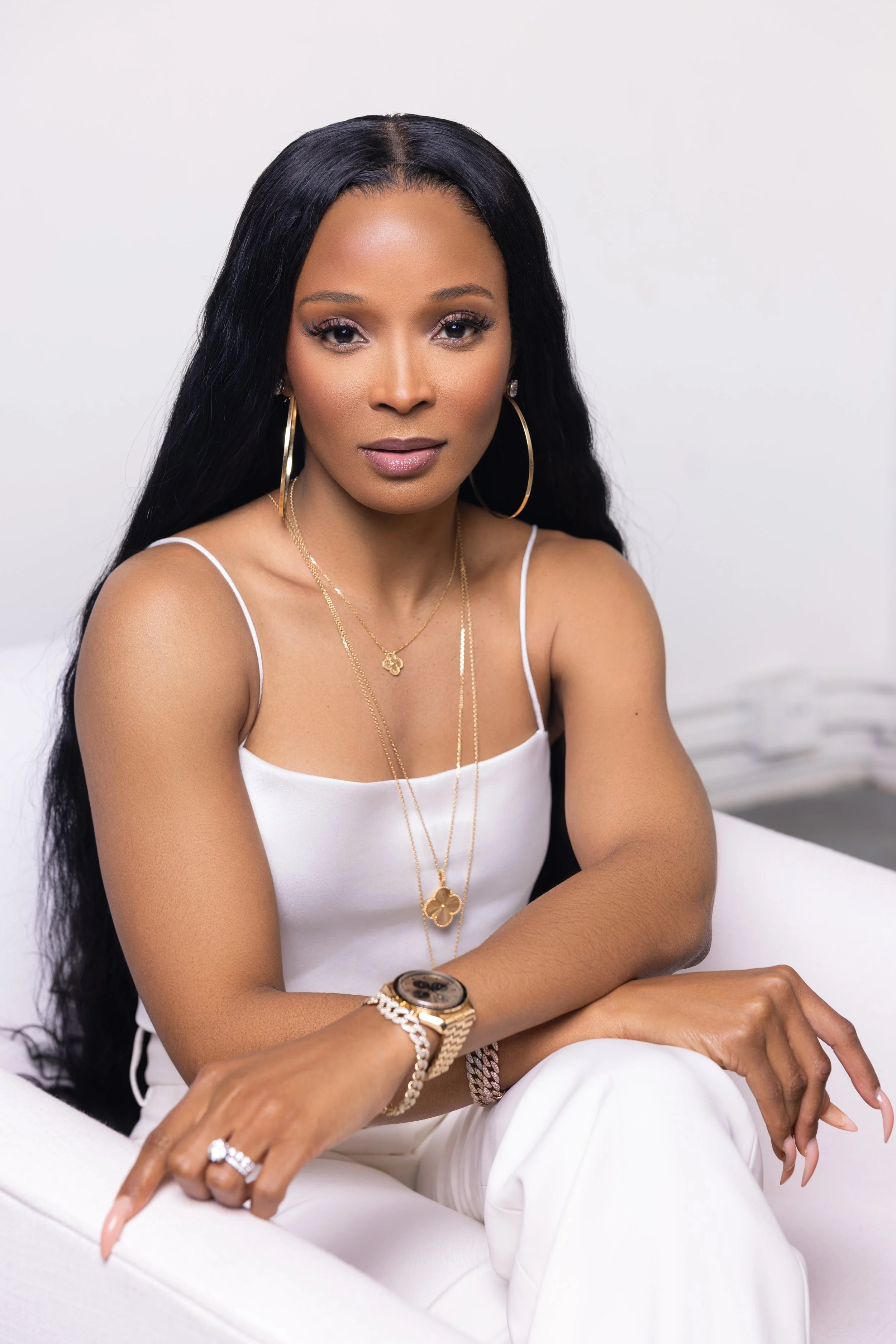 A woman with long black hair wearing a white tank top and gold jewelry, sitting on a white chair, looking at the camera.