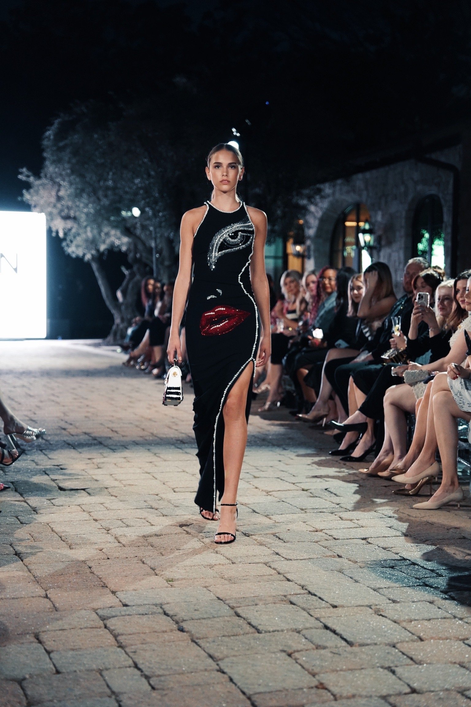A fashion model walks down the runway during a fashion show, wearing a black dress with artistic embroidery of an eye and lips. The audience is seated on both sides, watching the runway.
