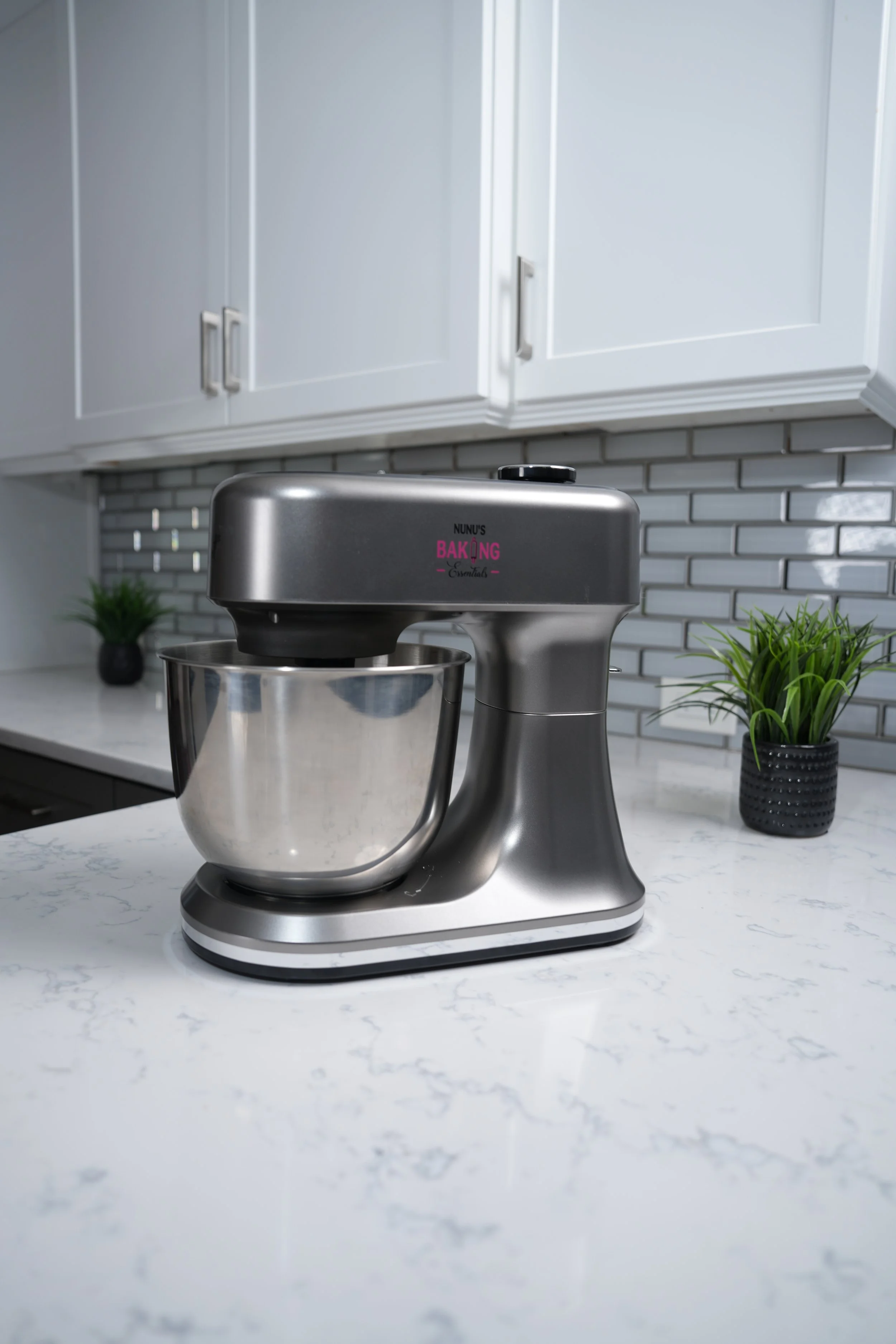 Stainless steel stand mixer on a white marble kitchen counter with cabinets, green plants, and a gray brick backsplash in the background.