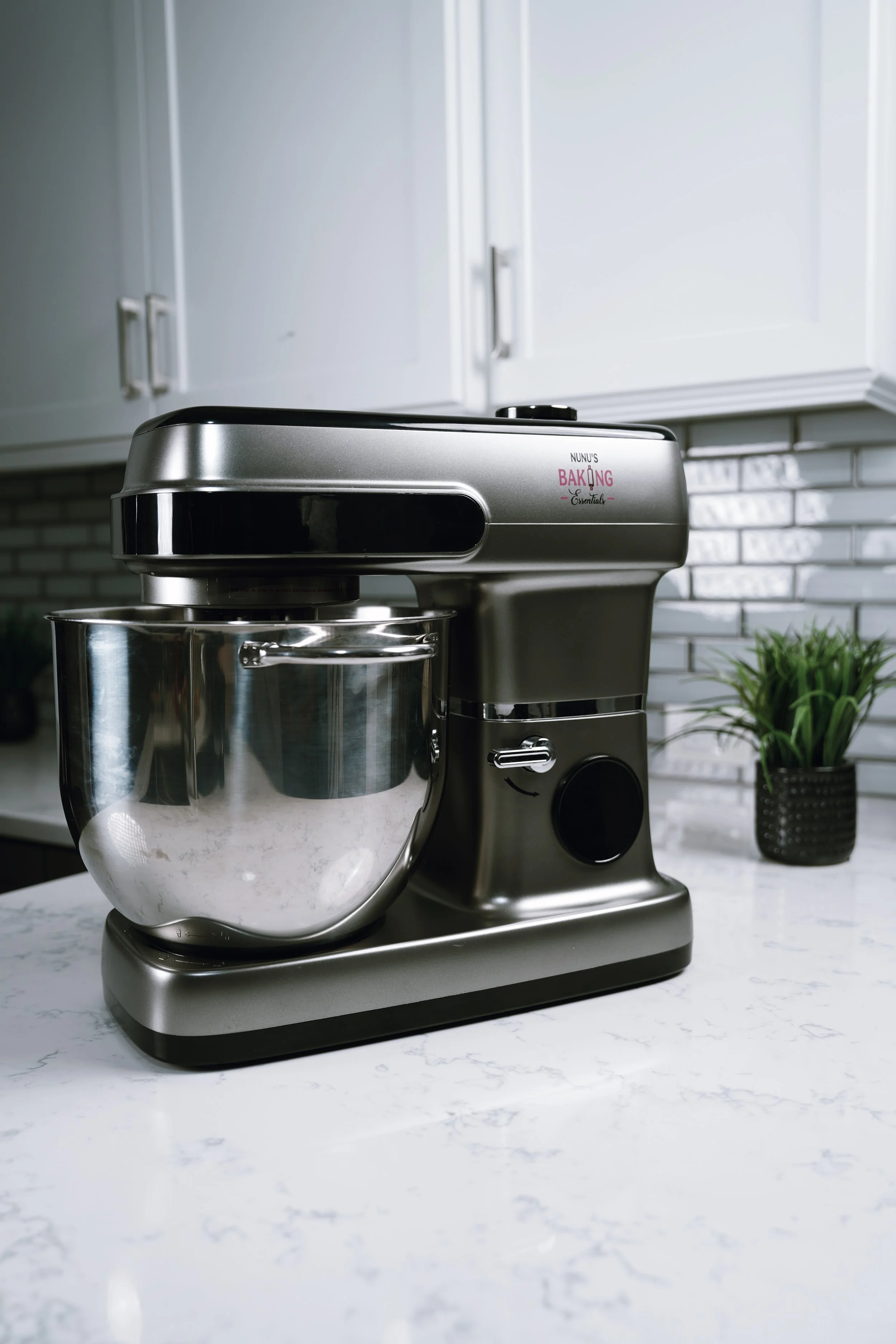 A silver kitchen stand mixer with a mixing bowl on a white marble countertop, with white cabinets and a small plant in a black pot in the background.