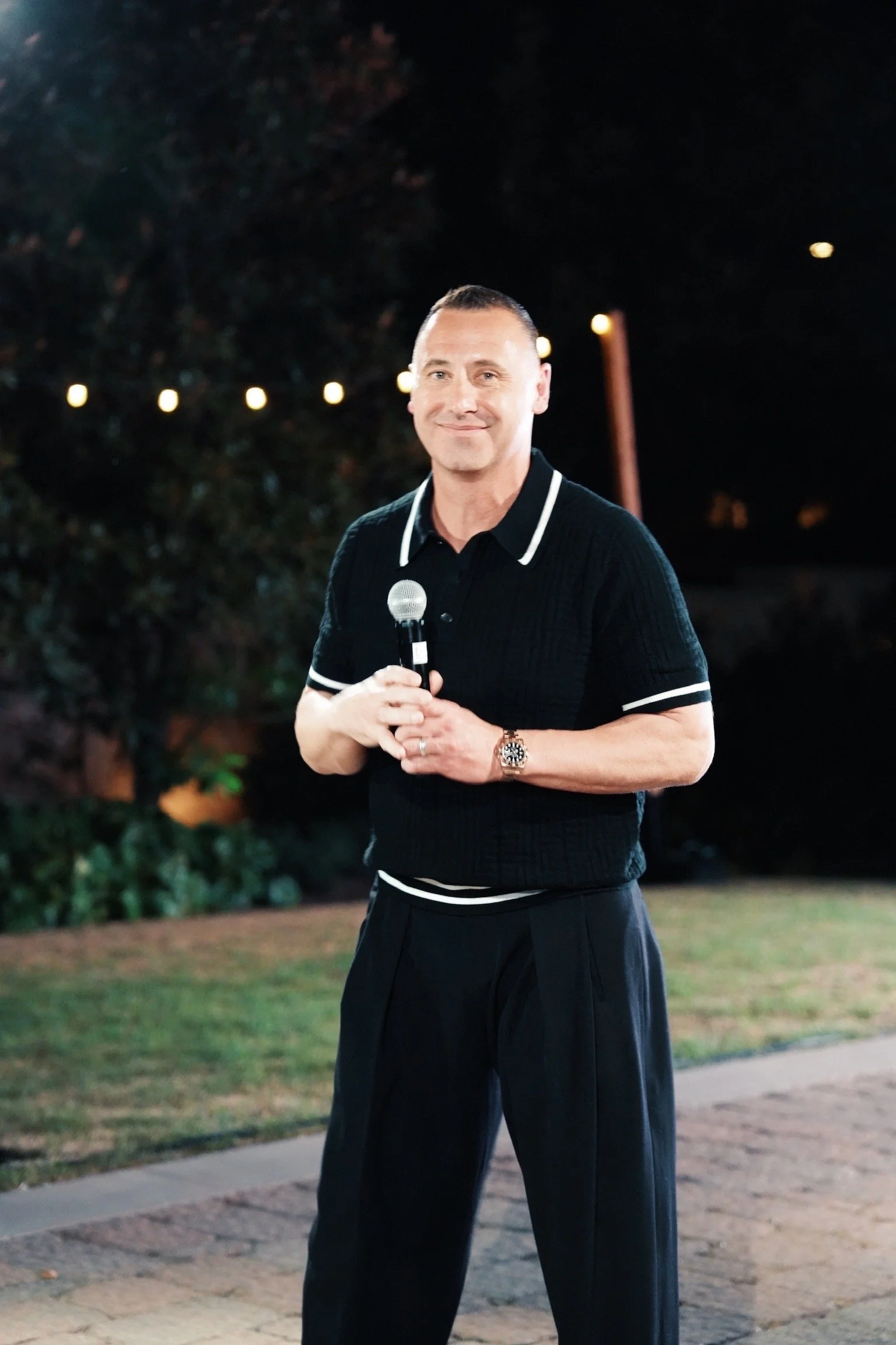 A man standing outdoors at night, holding a microphone, smiling, wearing a black polo shirt with white trim and black trousers, with blurred string lights in the background.