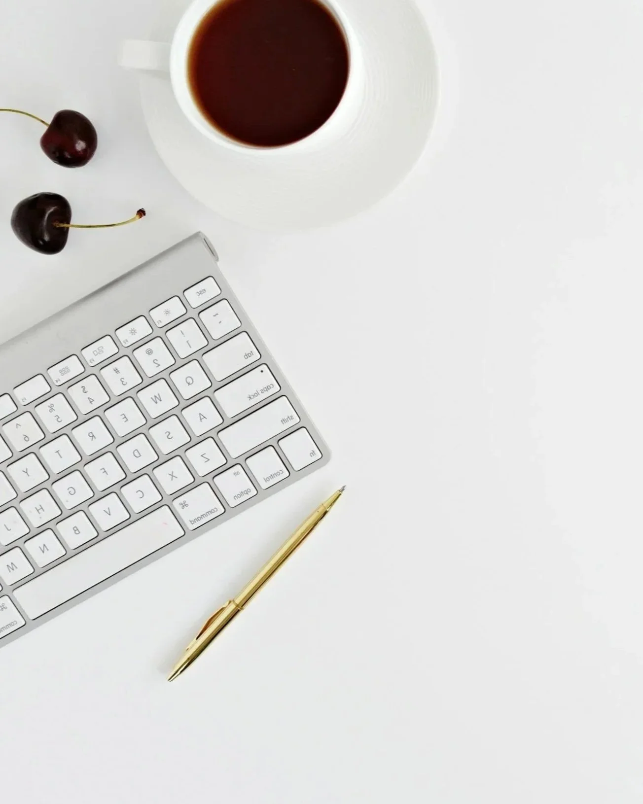 A white desk with a wireless keyboard, a gold pen, a cup of black coffee, and two cherries.