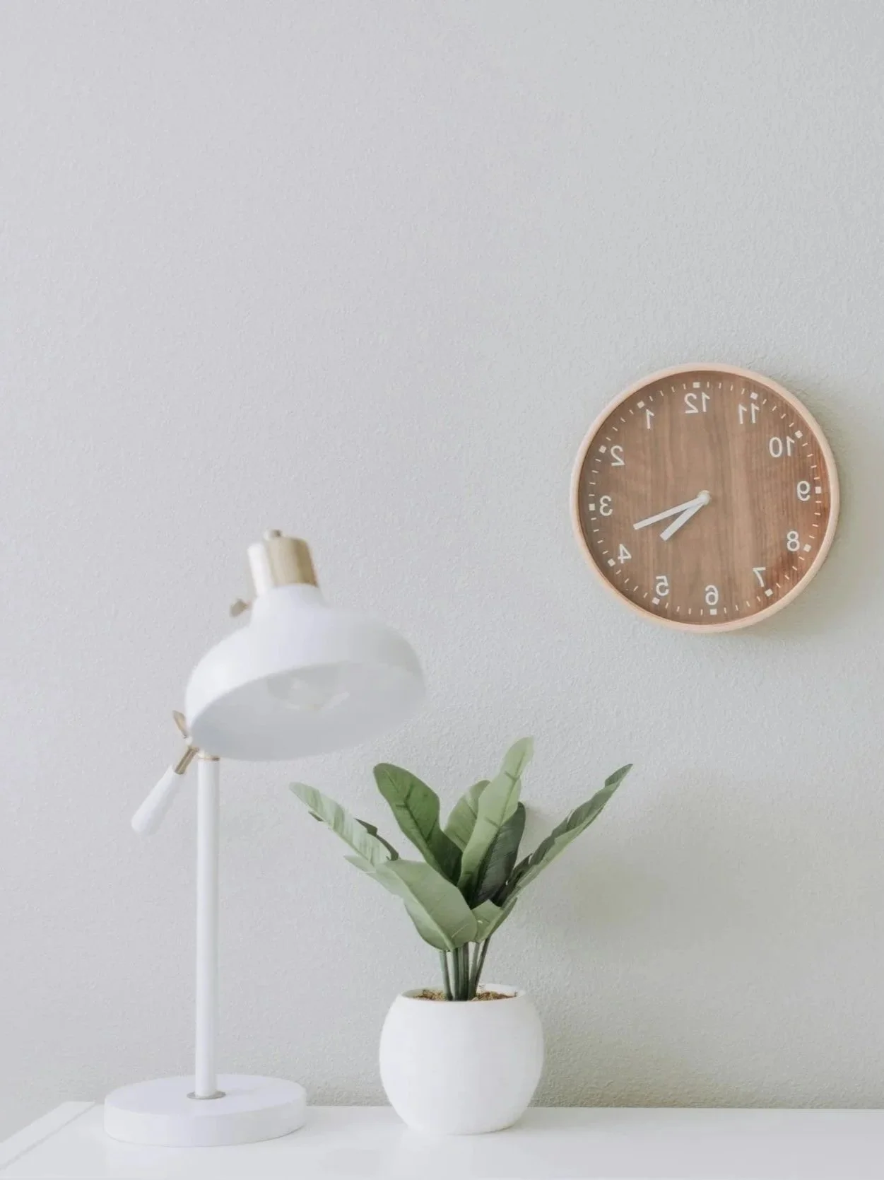 White desk lamp, potted green plant, and a round wall clock with a wooden face showing the time as 3:30, against a plain white wall.
