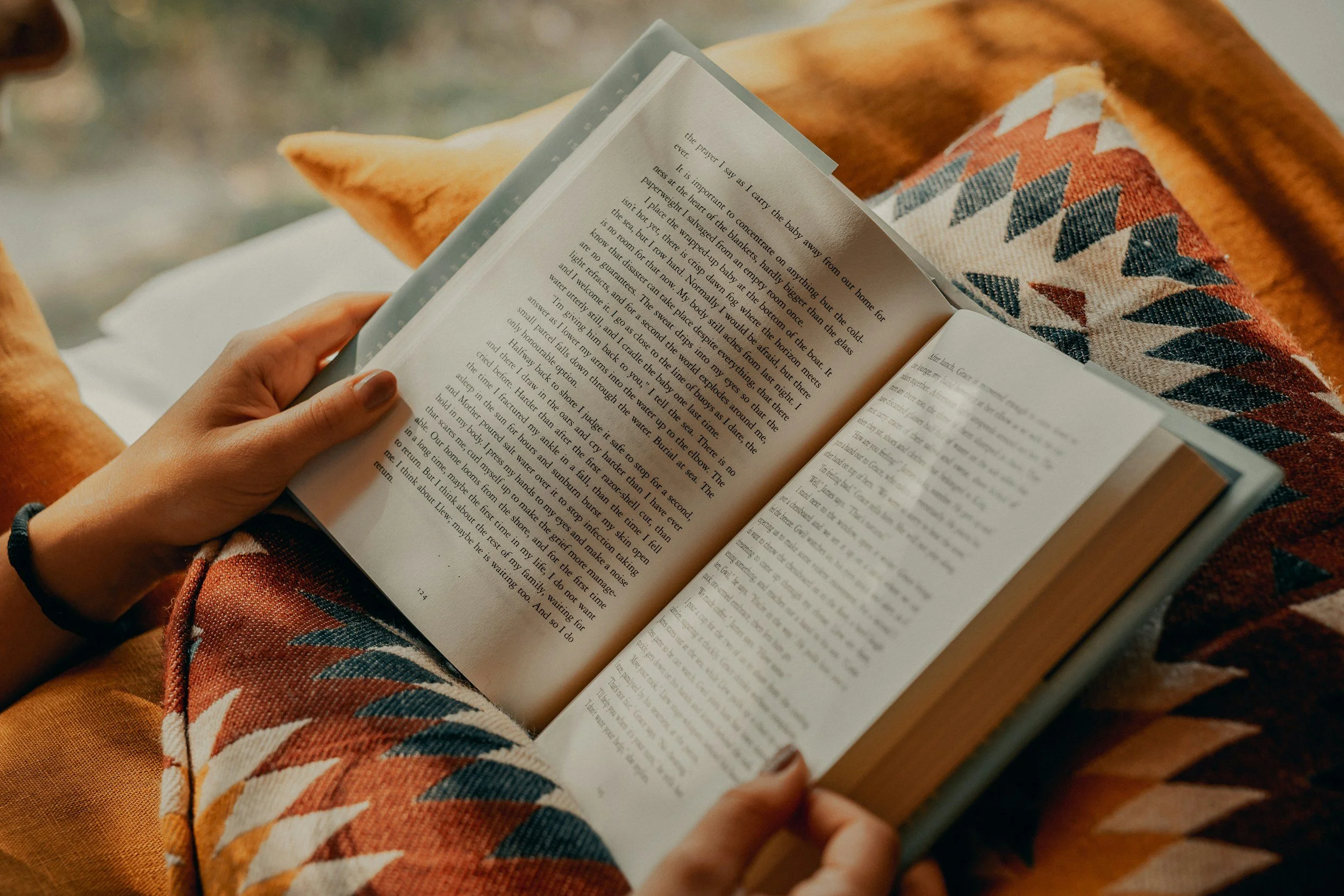 Close up on an open book resting on a pillow, being held by the hands of a teenager.