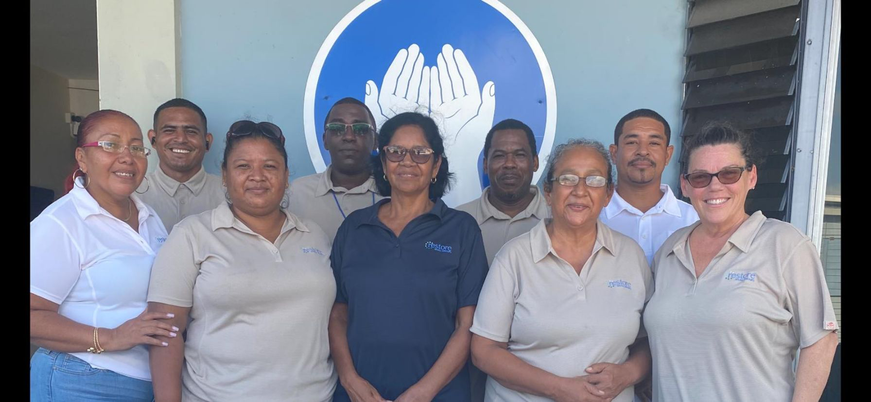 Group of nine people standing in front of a wall with a logo of two hands cupped together.