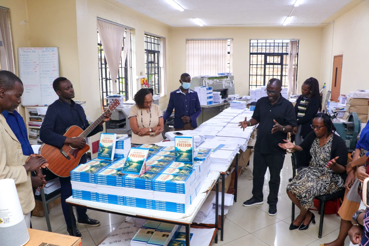 Group of people in an office, some standing and some sitting, around a table with stacks of books and papers. One person is playing a guitar, others are engaged in conversation or listening.