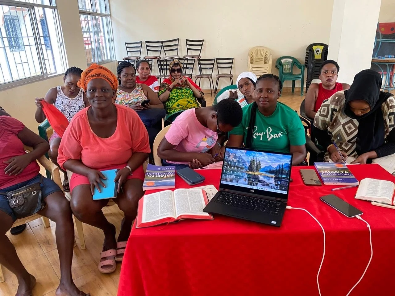 Group of women sitting around a red table with books, notebooks, a laptop, and phones in a room with windows and empty chairs in the background.