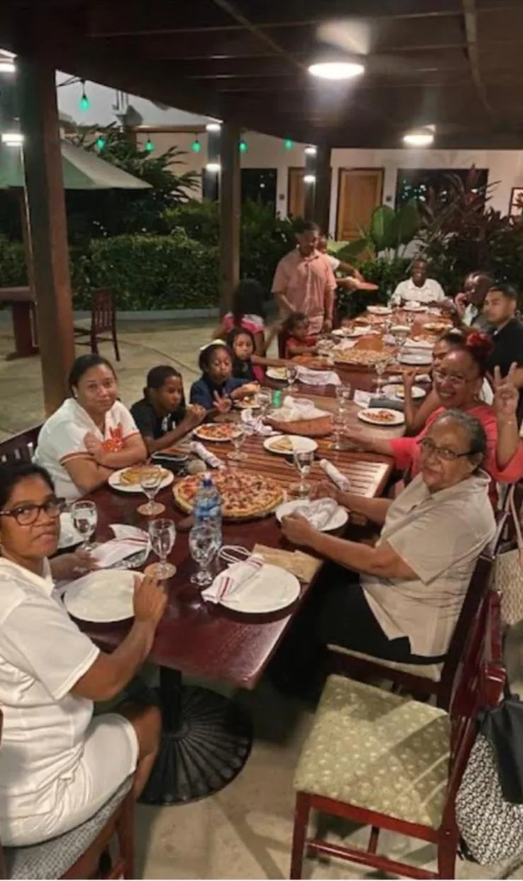 A large family gathering at a dinner table outdoors under a covered patio, with pizza, drinks, and children present