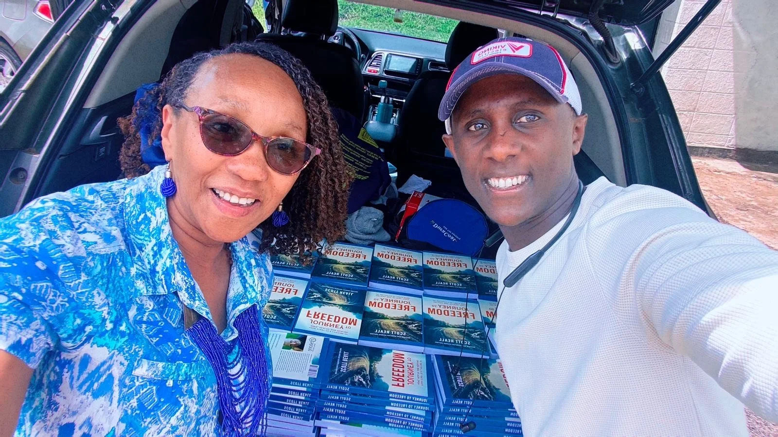 A woman and a man take a selfie in front of an open car trunk filled with copies of a book titled 'Reedom' by Joyce O. Scott. The woman wears sunglasses, a blue patterned shirt, and blue jewelry. The man is wearing a white shirt, a cap, and has a headset around his neck.