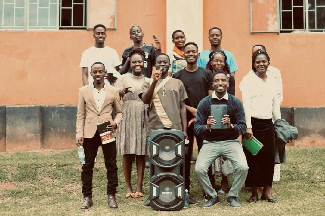 Group of eleven young adults standing outdoors in front of a peach-colored building with windows, smiling and posing for the photograph, with a large speaker and some books and water bottle in the foreground.