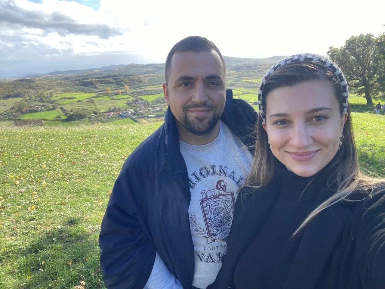 A man and woman taking a selfie outdoors on a grassy hill with a scenic landscape of mountains, fields, and trees in the background.
