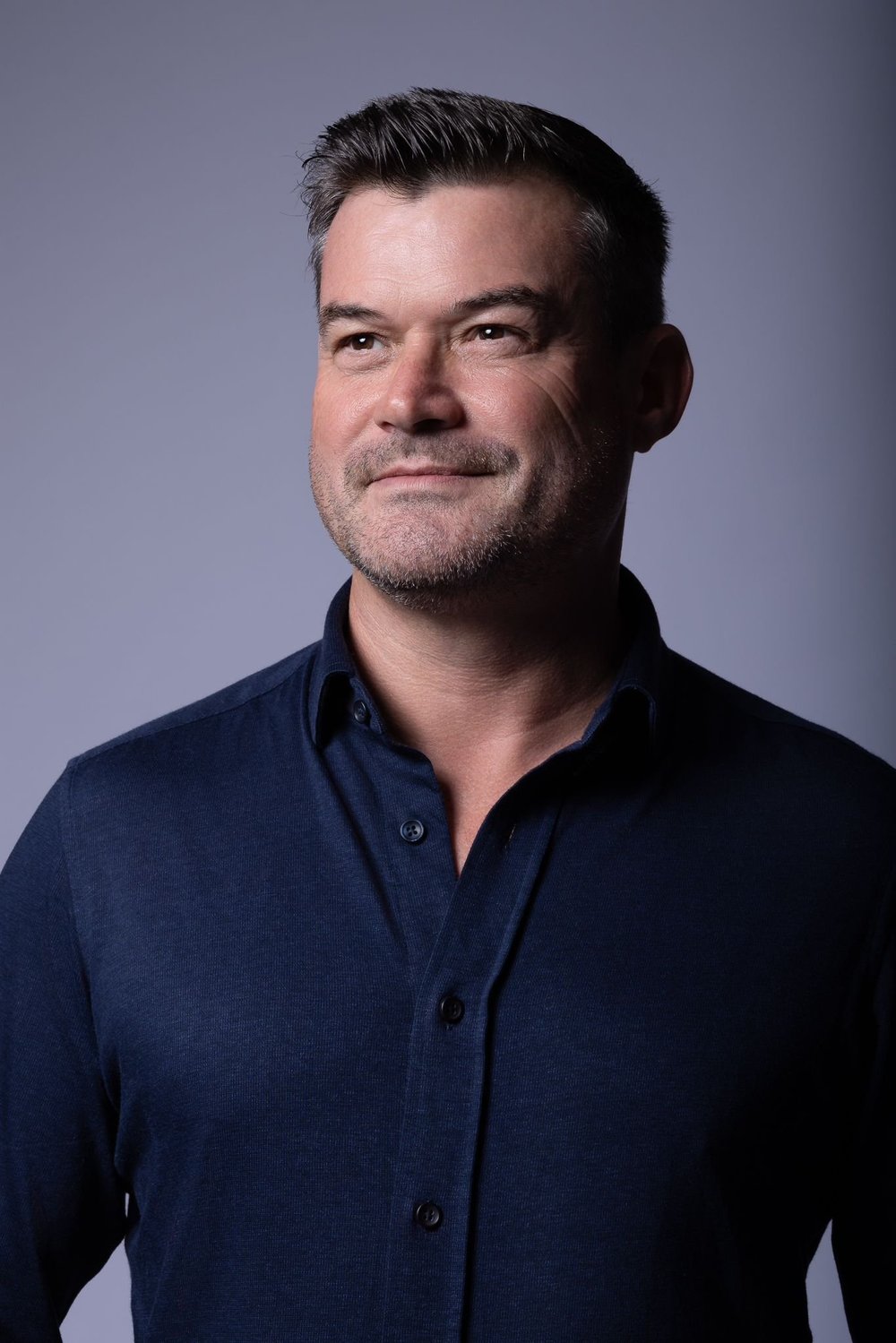 Portrait of a middle-aged man with short dark hair and light beard, wearing a dark blue button-down shirt, looking slightly to his right against a plain gray background.