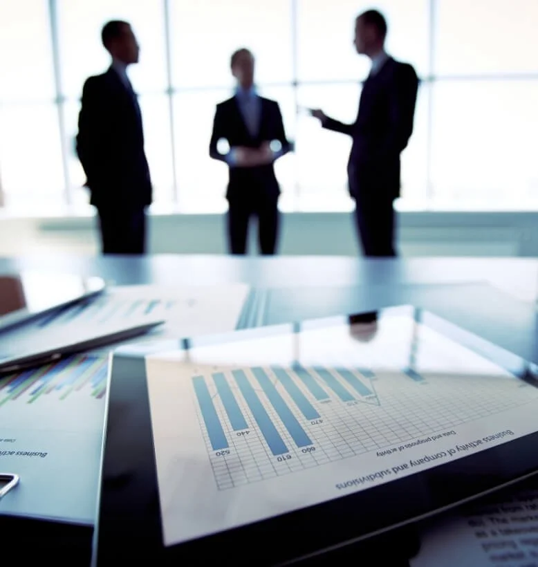 Silhouettes of three business professionals standing and talking in a bright office with large windows, with a table in the foreground featuring business documents and charts.