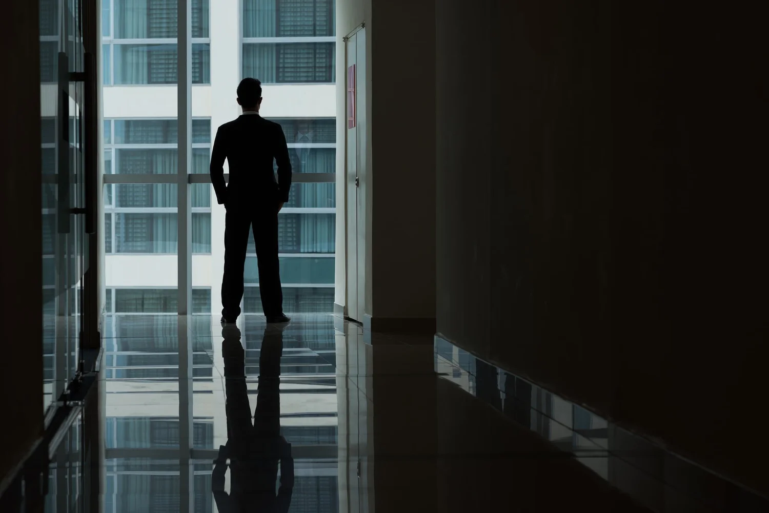 A silhouette of a man in a suit standing in a hallway, looking out a large window at a modern office building.