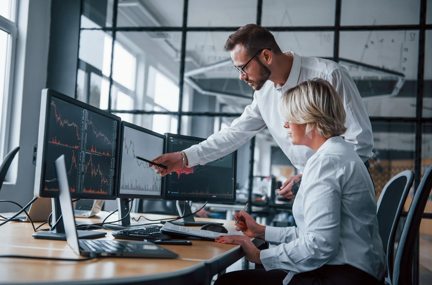 Two professionals working with multiple computer monitors displaying stock charts in a modern office.