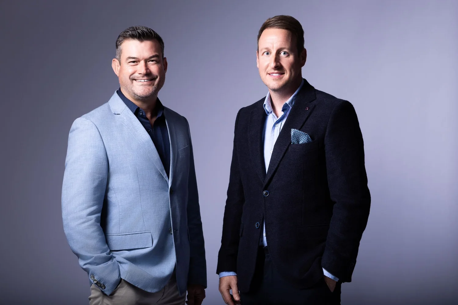 Two men in business suits standing against a gray background, smiling at the camera.