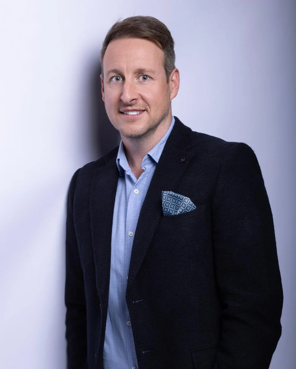 A man with short brown hair wearing a light blue collared shirt and a black blazer with a patterned pocket square, standing against a plain light gray background.