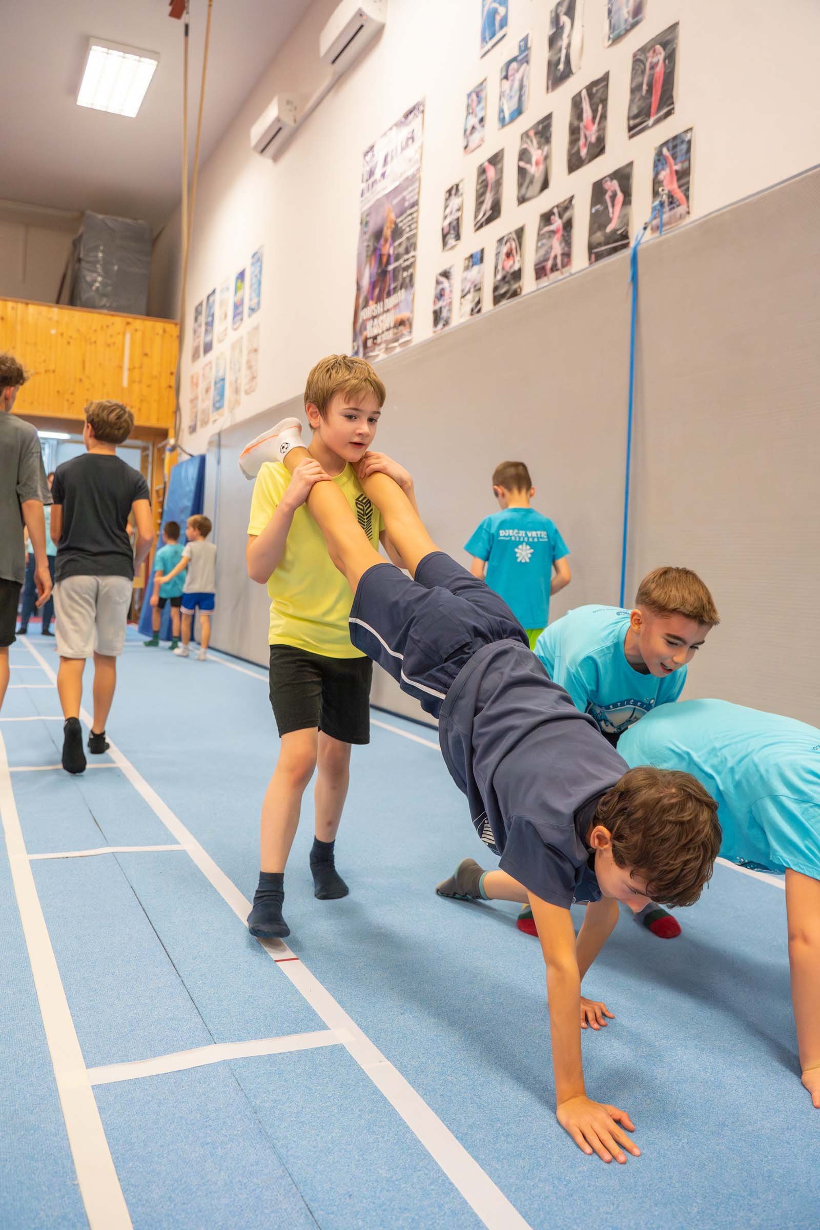 Djeca u gimnastičkoj dvorani, jedno izvodi vježbu dok drugo drži njegove noge, okruženi ostalom djecom koja hodaju.