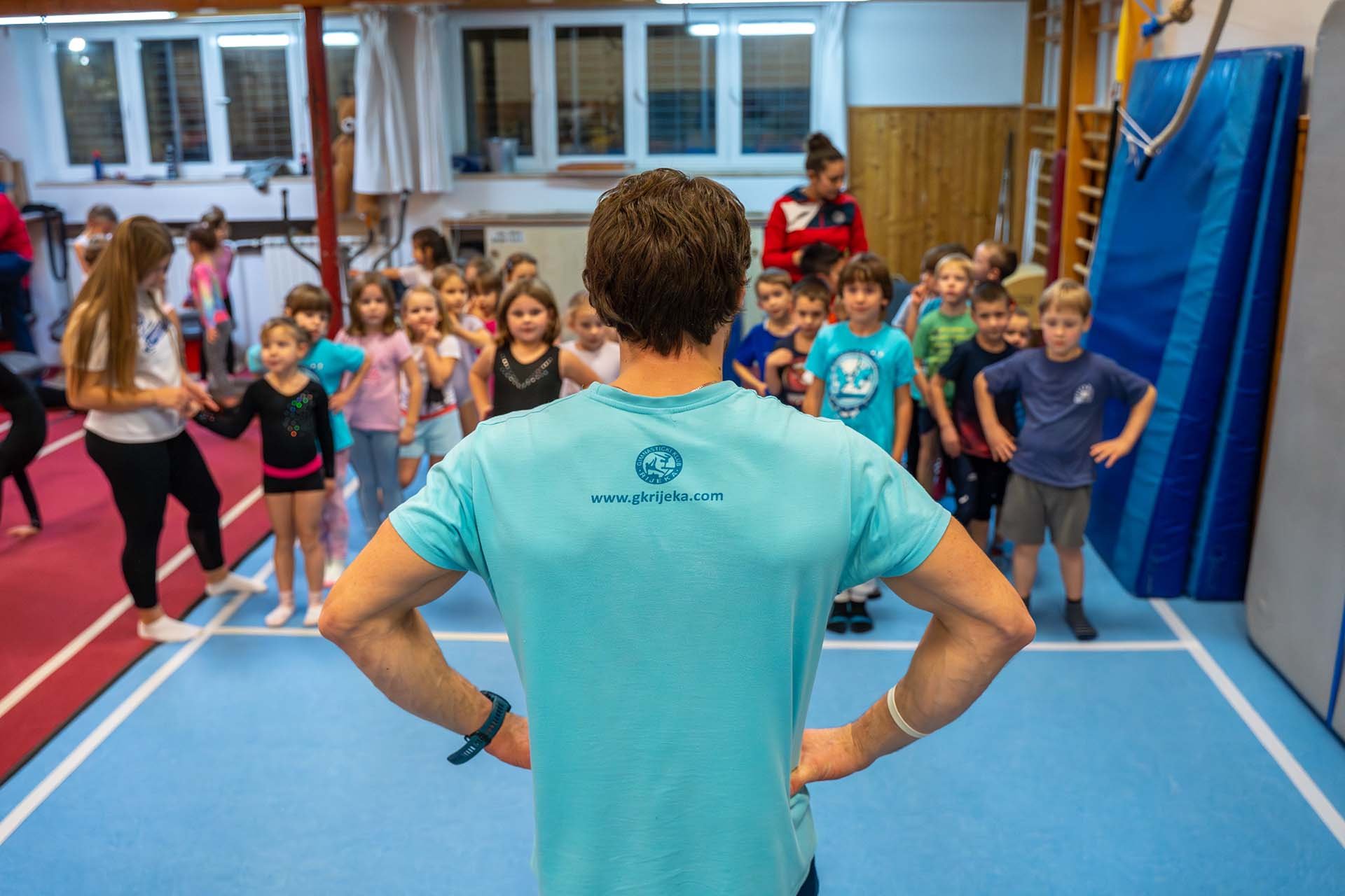 Učitelj gimnastike okrenut prema grupi djece u gimnastičkoj dvorani.
