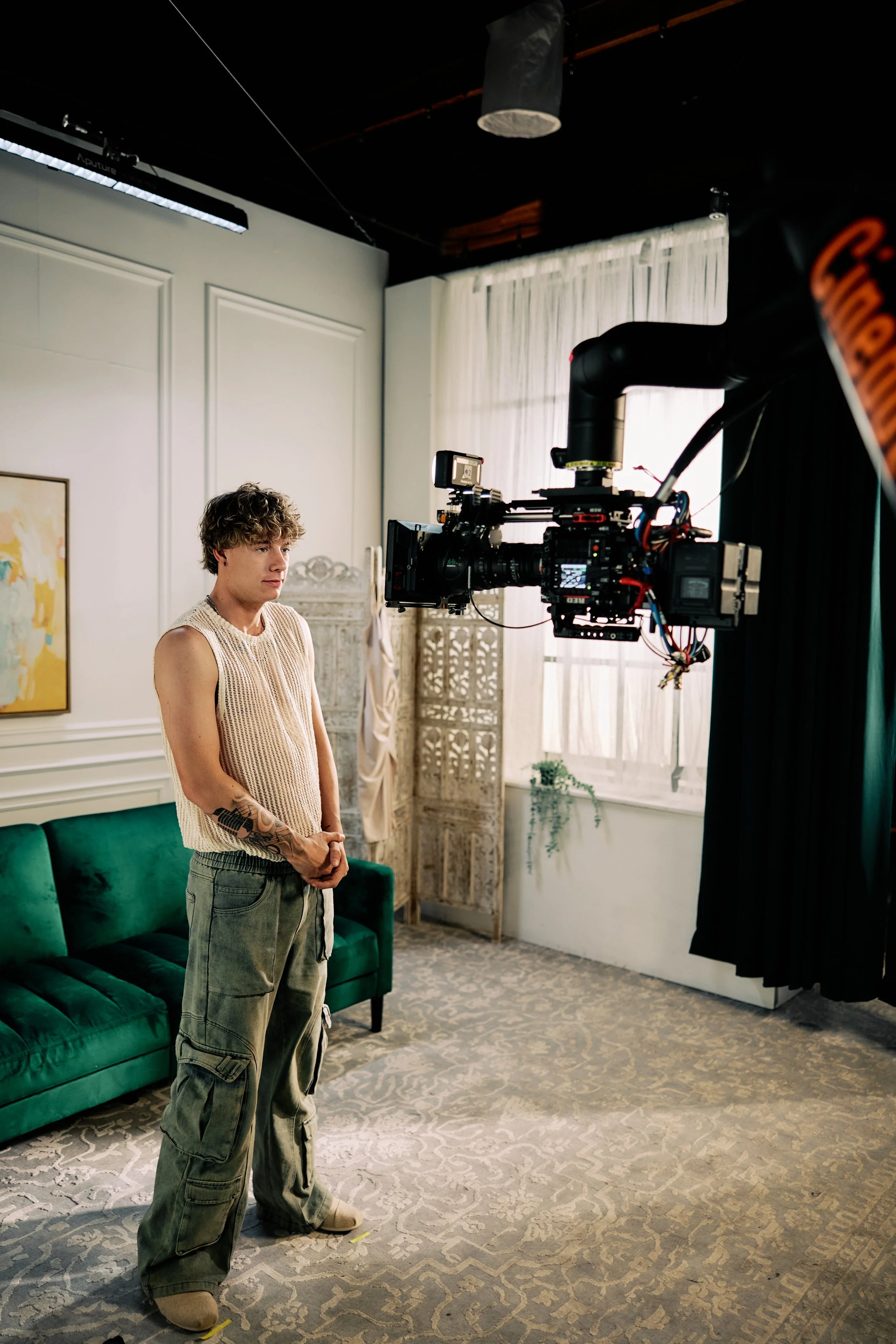 Performer standing beside a robot-mounted cinema camera on an interior set with a green sofa.