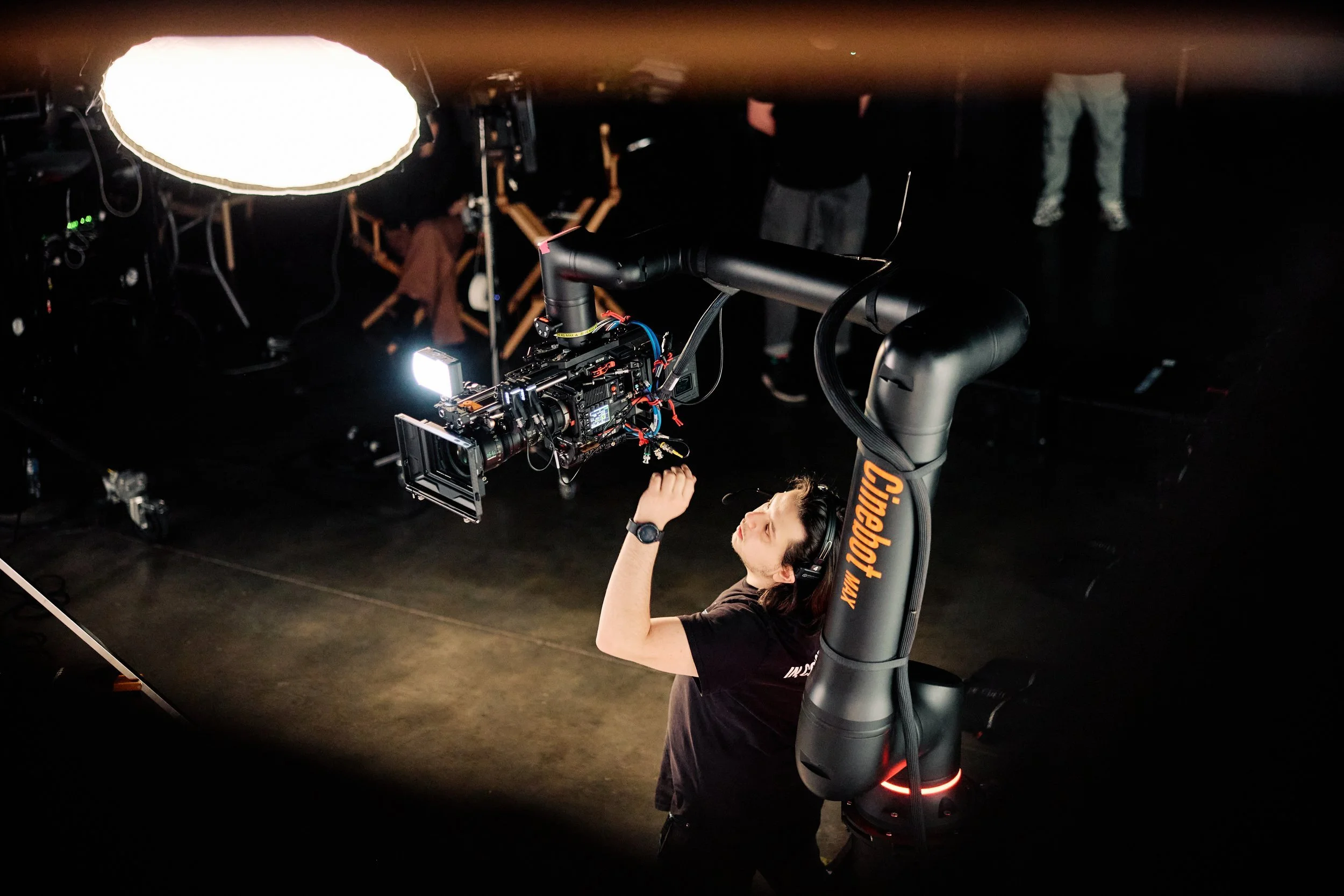 Crew member reaching toward a cinema camera mounted on the Cinebot Max arm beneath set lighting.