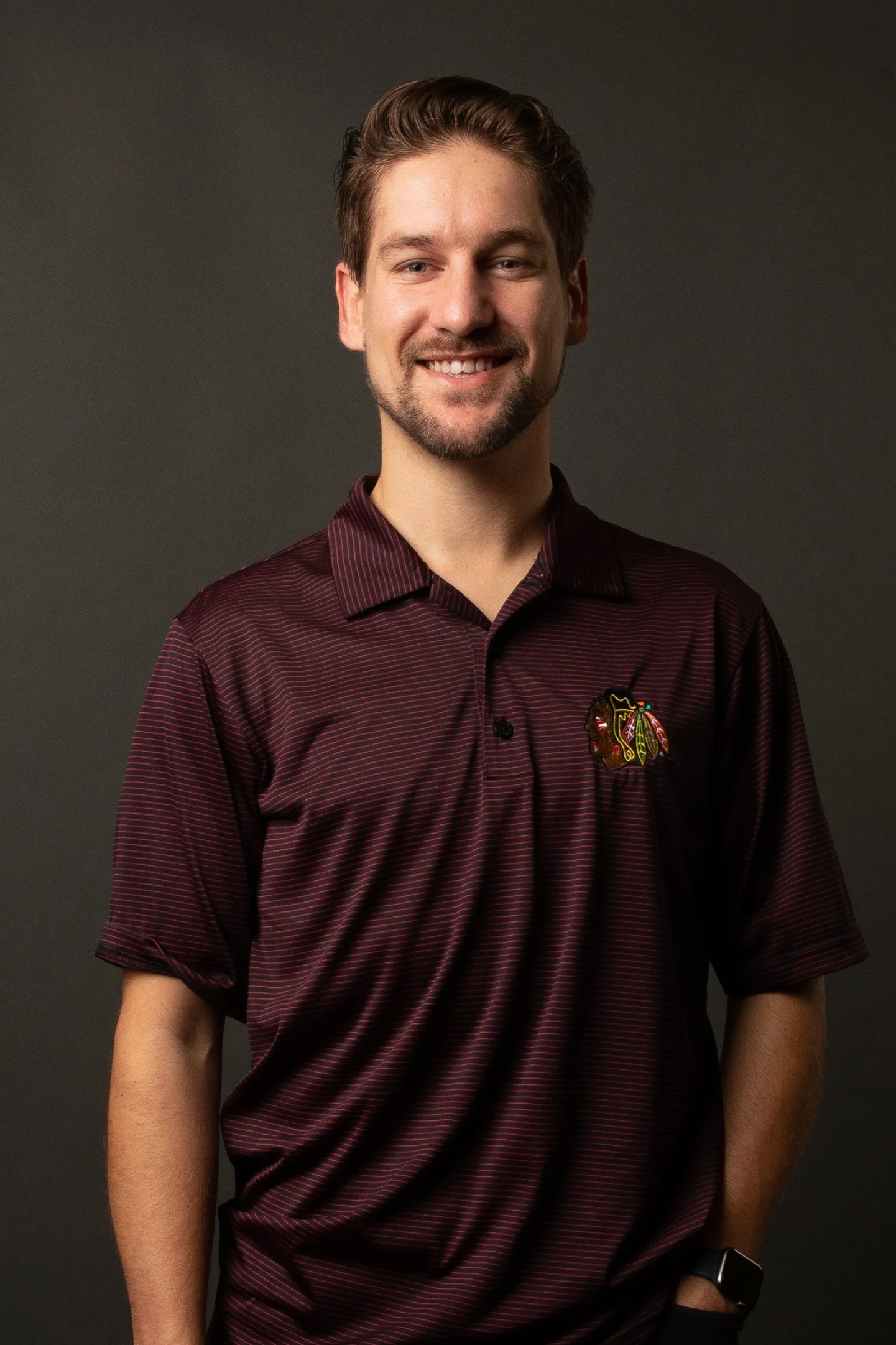 A smiling young man with light skin, brown hair, and a beard, wearing a maroon Chicago Blackhawks polo shirt with a logo on the chest, standing against a dark background.