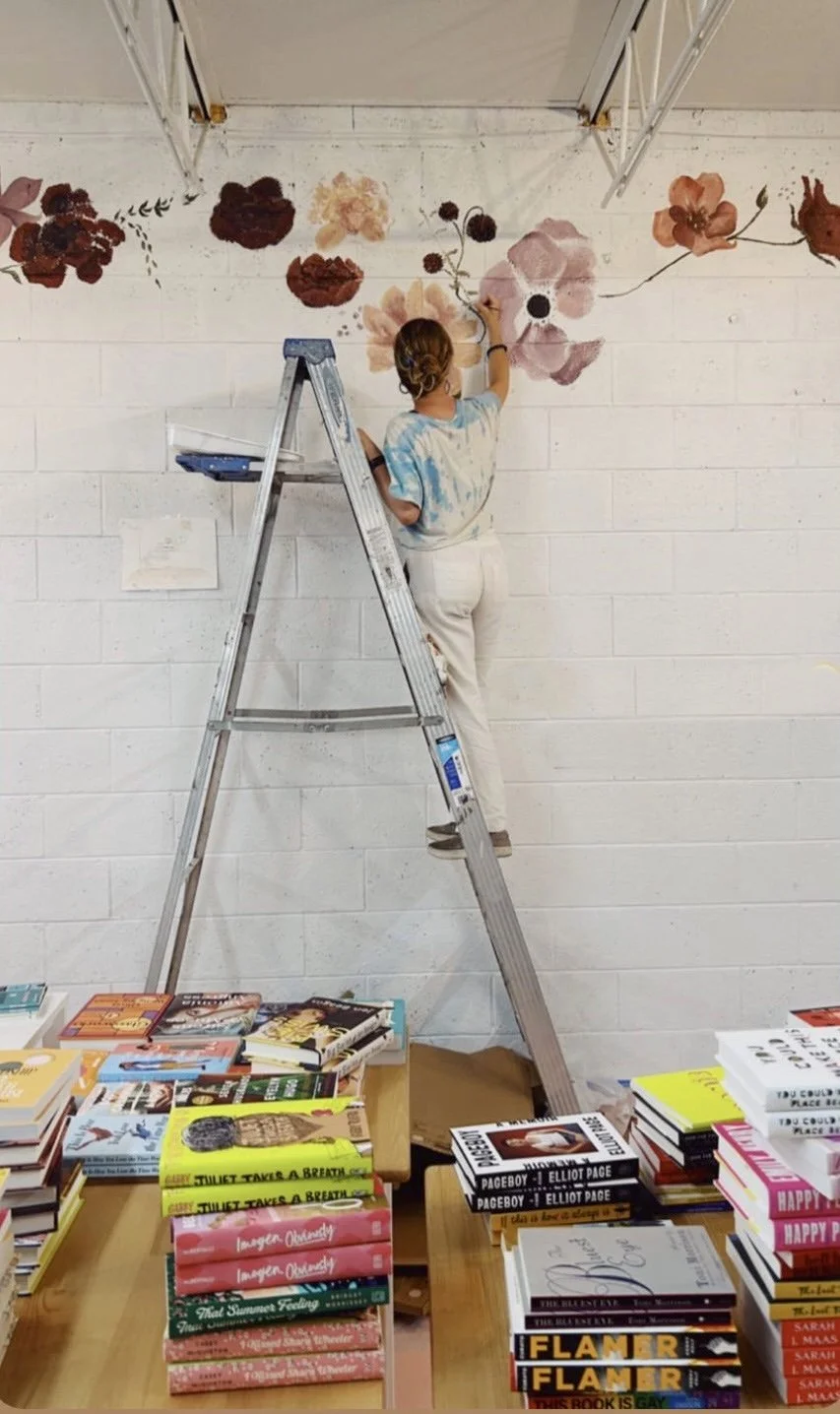 A person standing on a ladder painting a floral design on a white brick wall. The table below has stacks of colorful books.