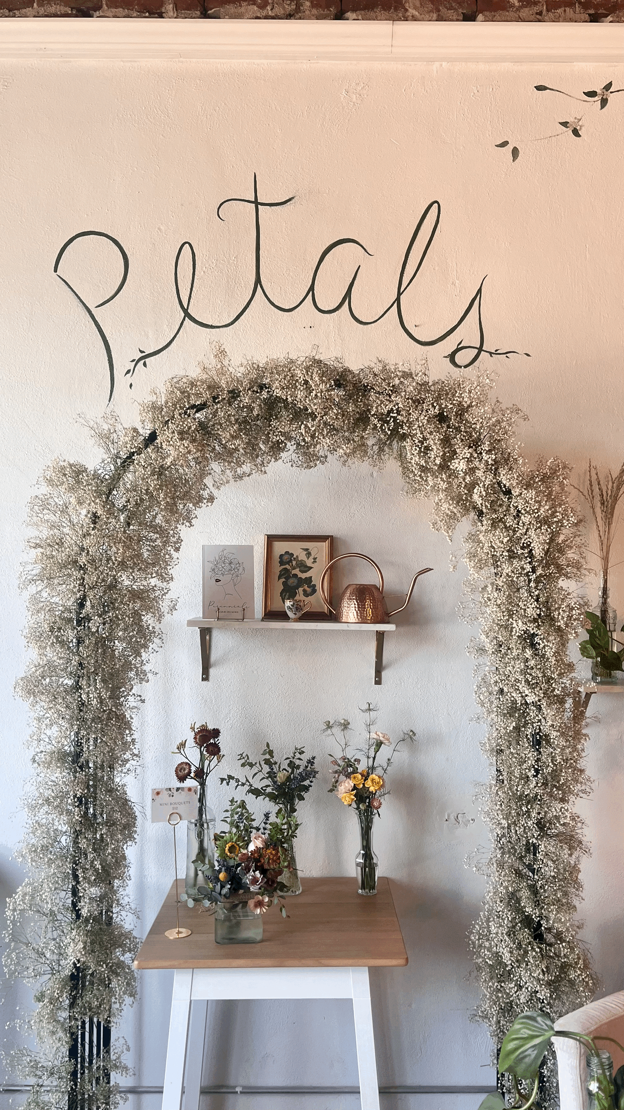 Decorative floral display with a white wall background that says 'Partly'. A white shelf holds a framed art piece, a framed photograph, and a copper teapot. On a wooden table, there are three vases with various flowers, and a small sign that reads 'M