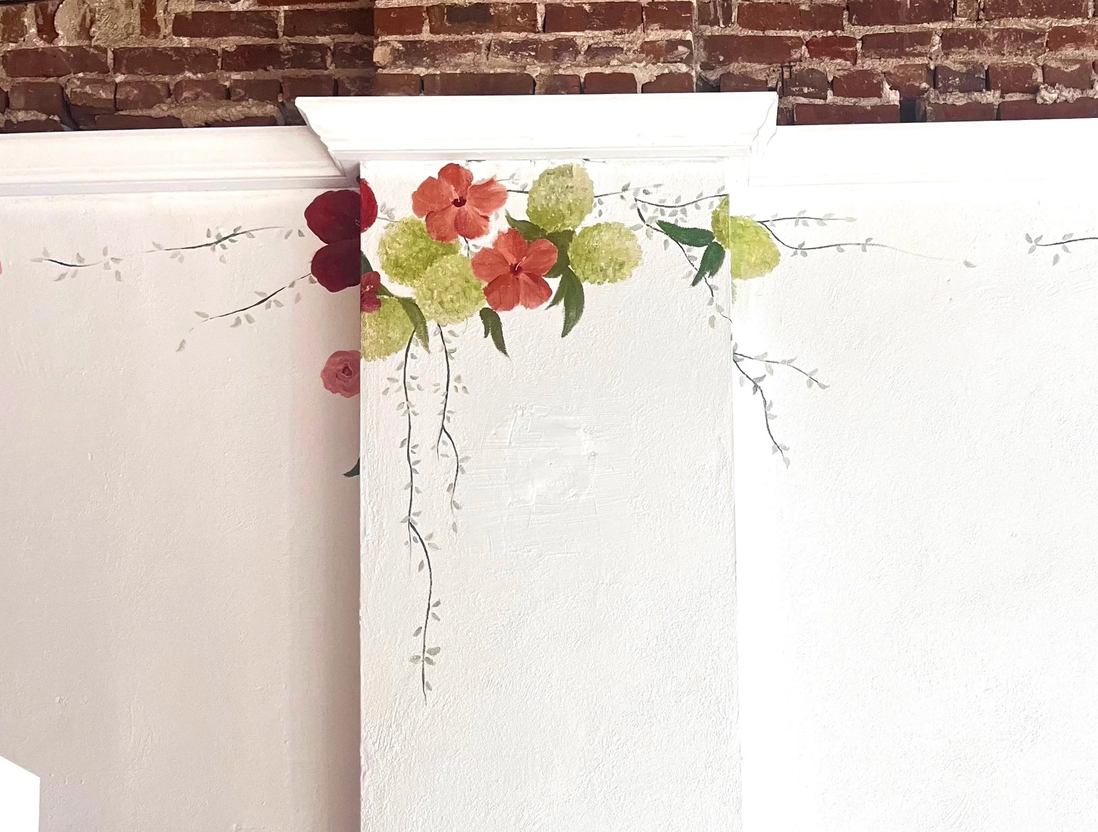 Decorative wall with painted flowers and vines on a white wall against a brick background.