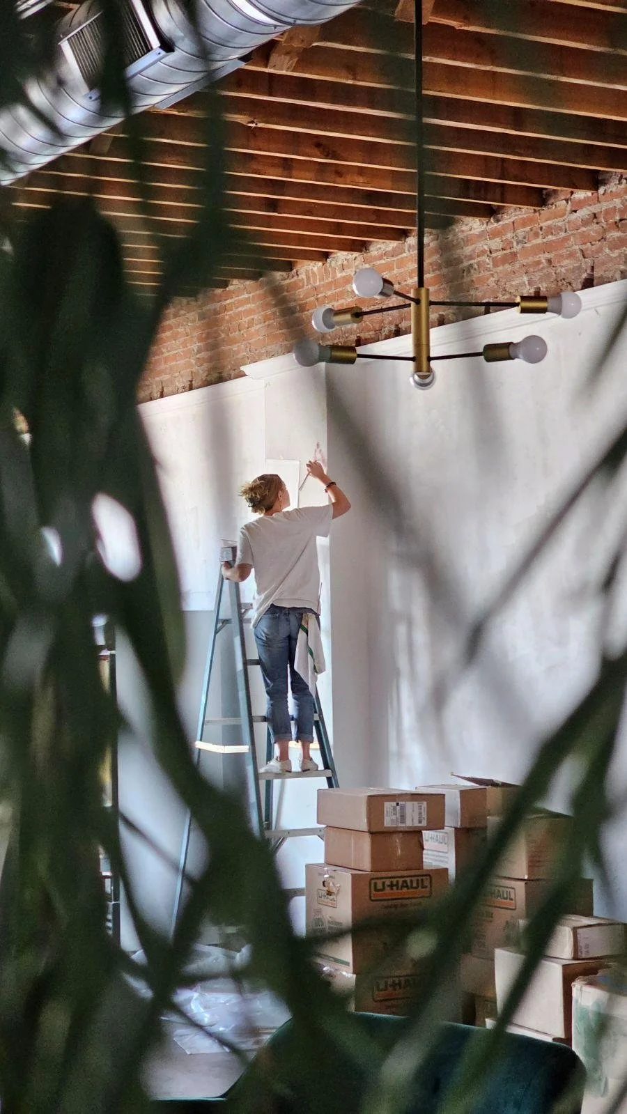 A woman standing on a ladder painting a white wall in a room with exposed brick ceiling and a modern chandelier, with moving boxes around.