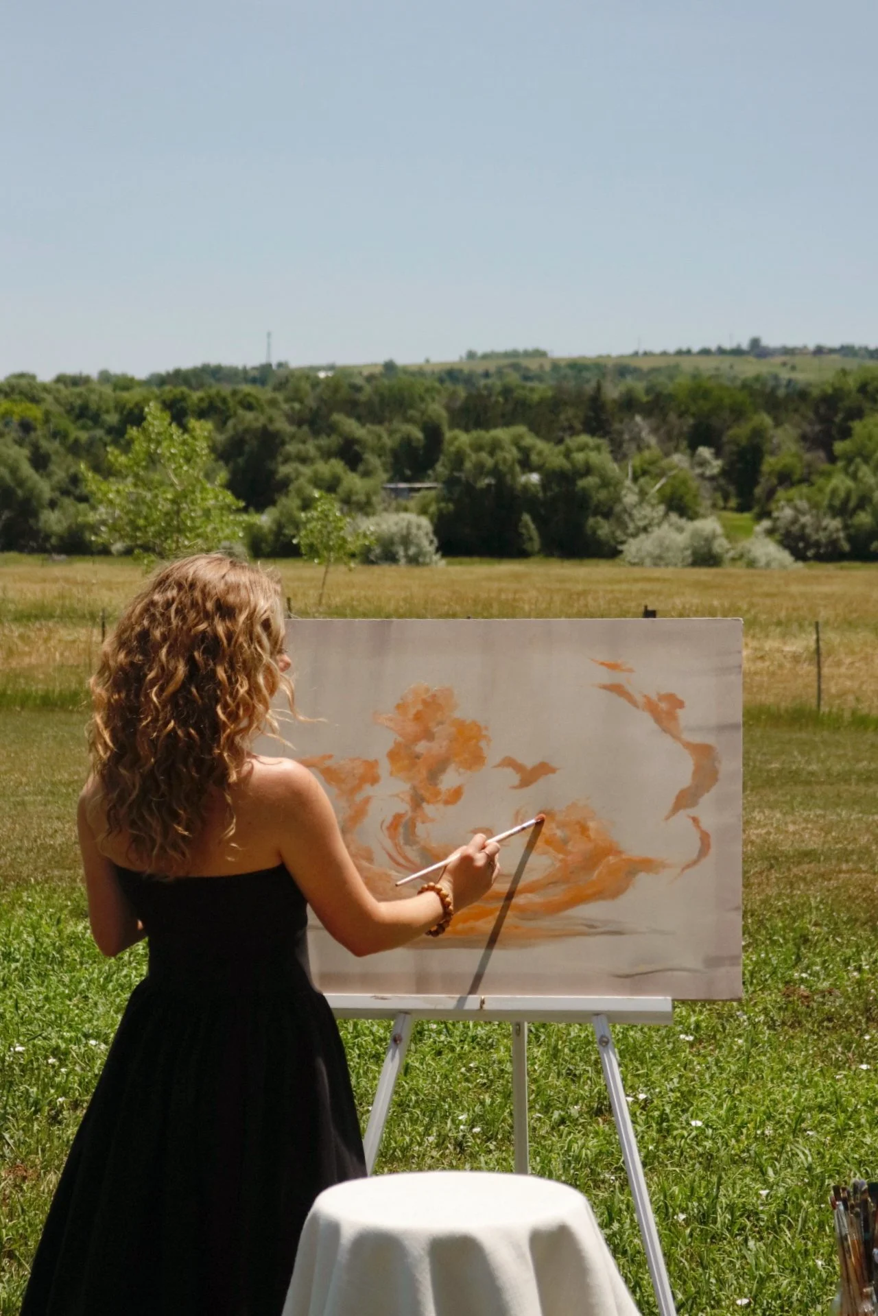 Artist Emma May Beers is painting on a large canvas outdoors, with a landscape of green fields and trees in the background.