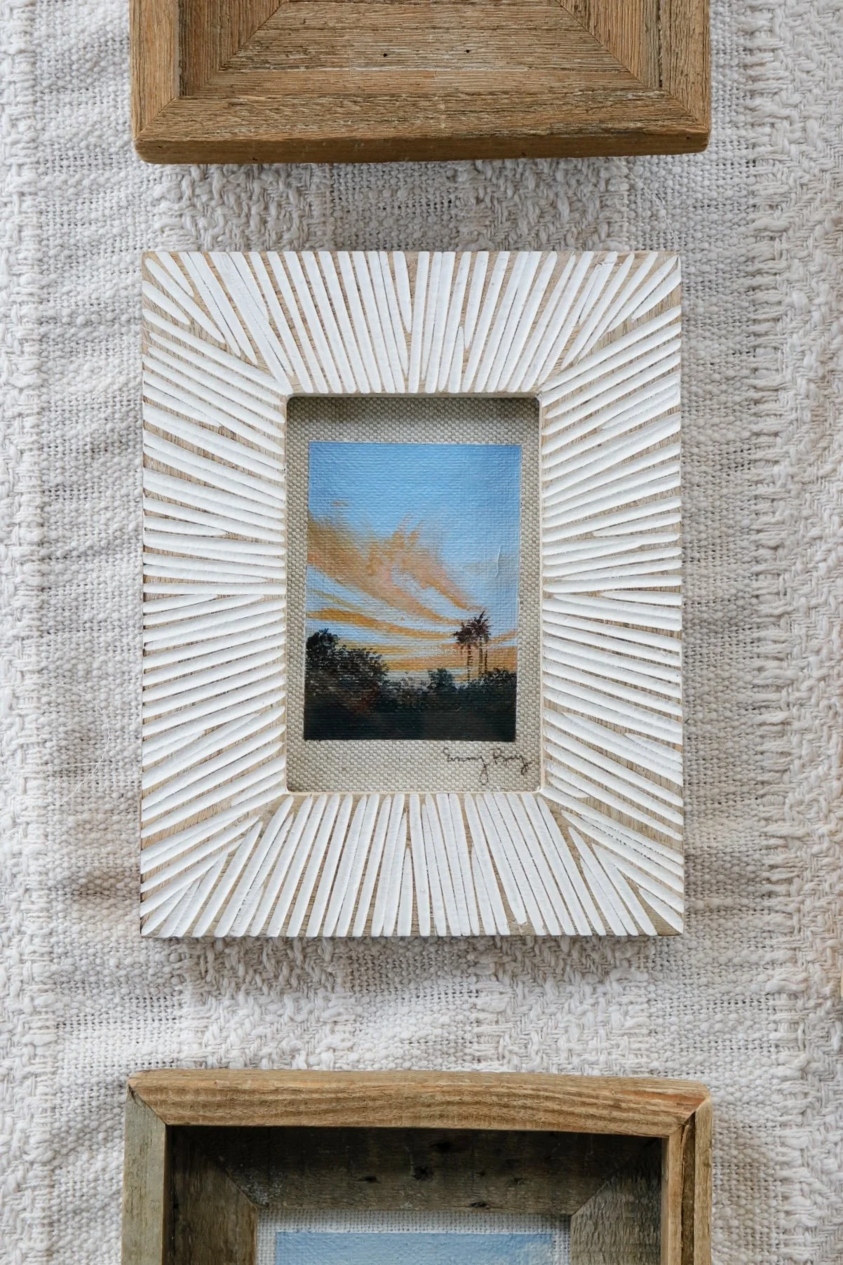 Miniature landscape painting of a sunset sky with orange clouds over dark trees, framed by a white, textured, sunburst-patterned frame with angled rays.