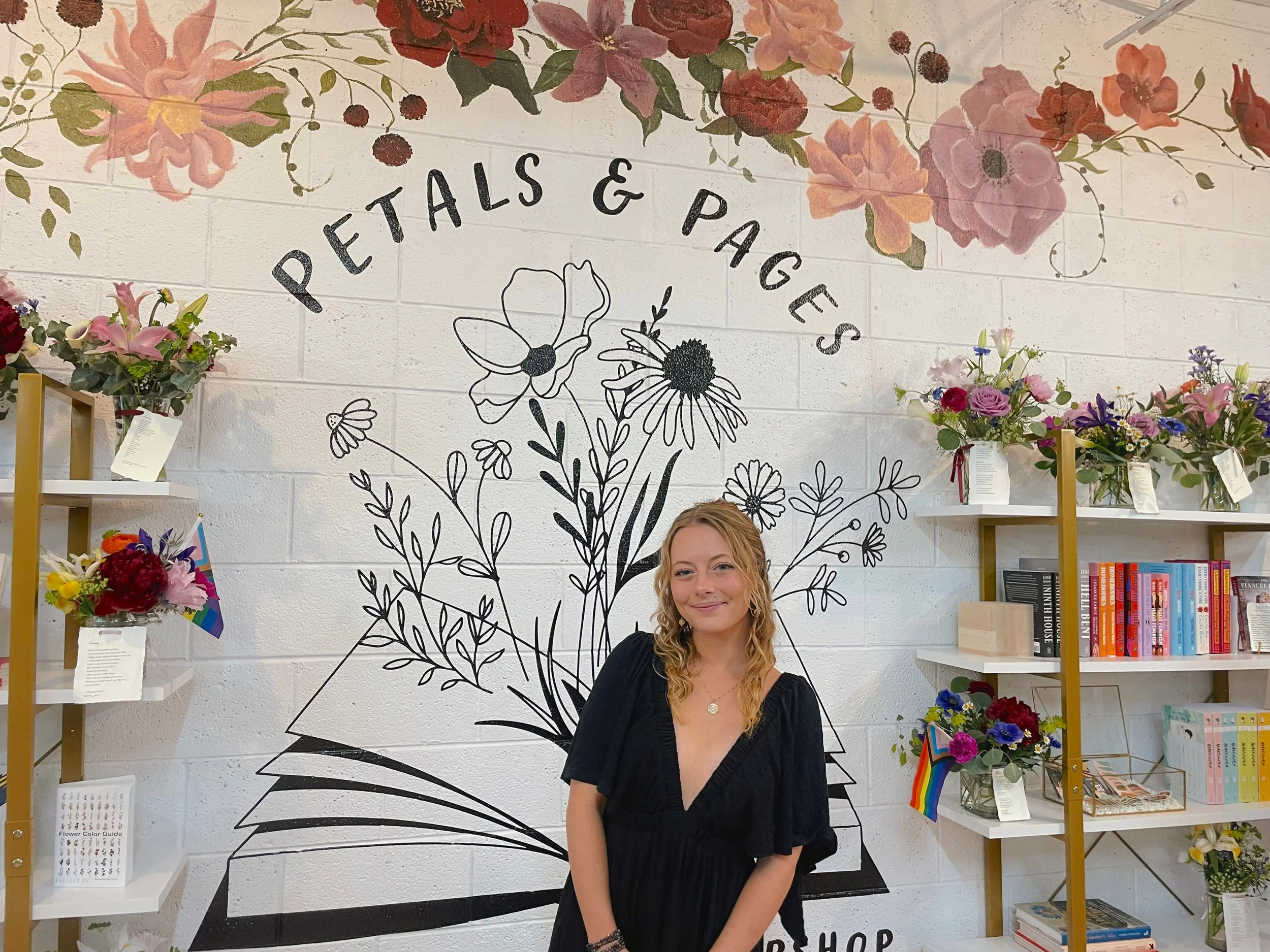 A young woman with blonde curly hair stands in front of a wall with forest floral artwork and the words 'Petals & Pages'. She is in a bookstore or flower shop, surrounded by shelves holding colorful books and flower arrangements.