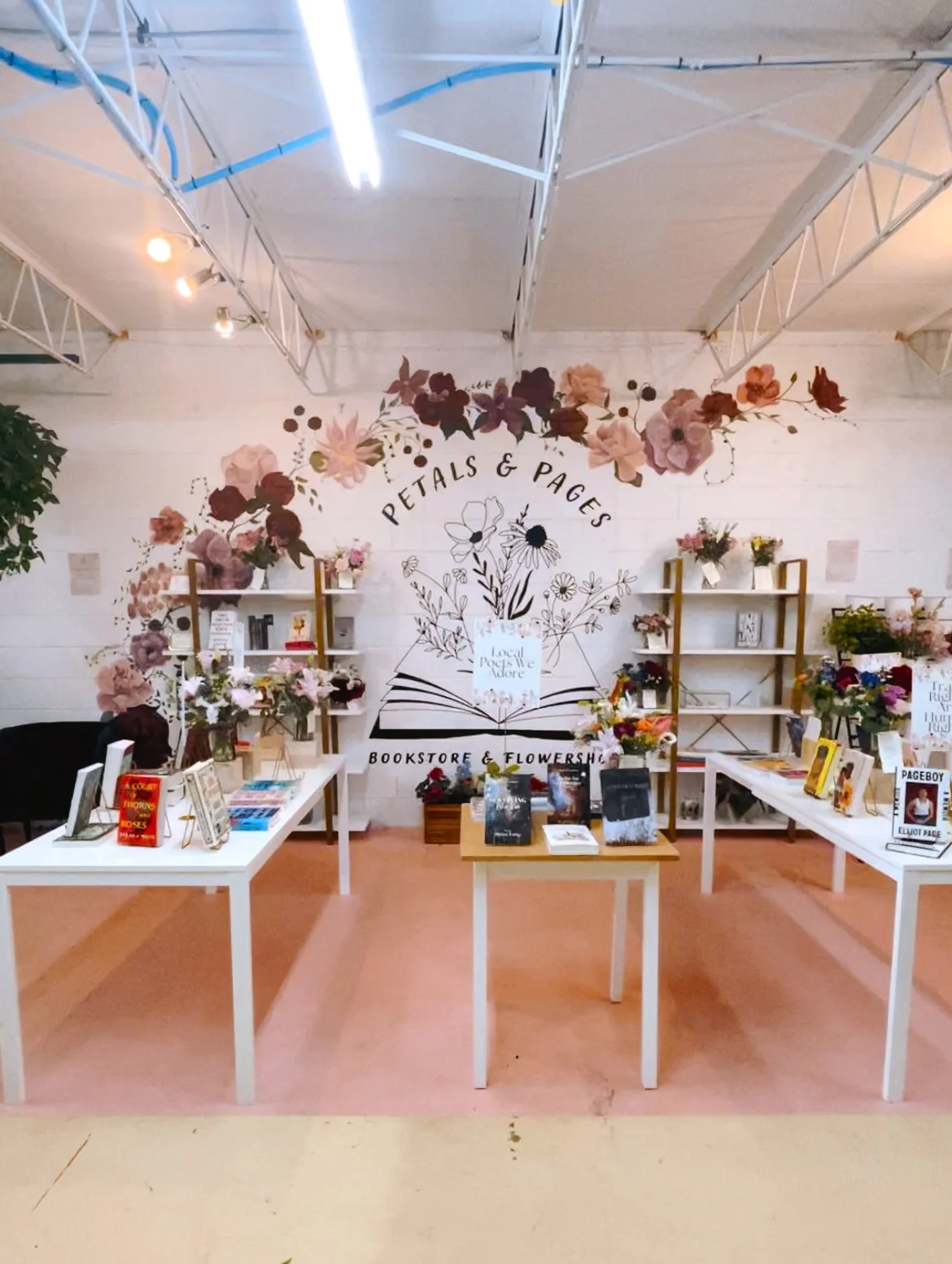 A bookstore and flower shop interior with a wall mural that reads 'Petals & Pages', decorated with floral artwork, books, and flowers displayed on white tables and shelves.