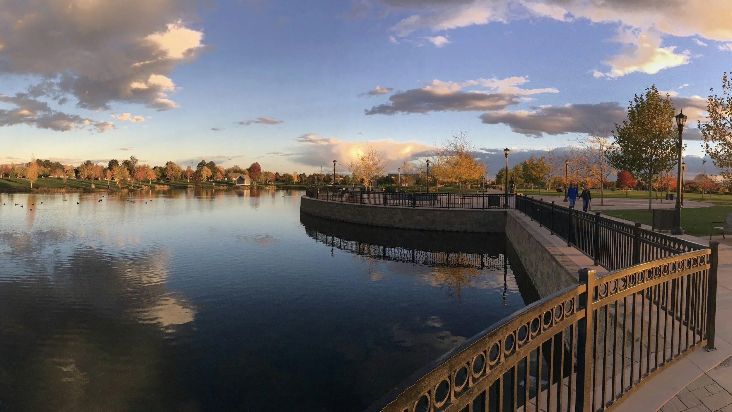 Scenic view of a lake with a park, trees with fall foliage, and a walking path with two people, under a sky with clouds during sunset.