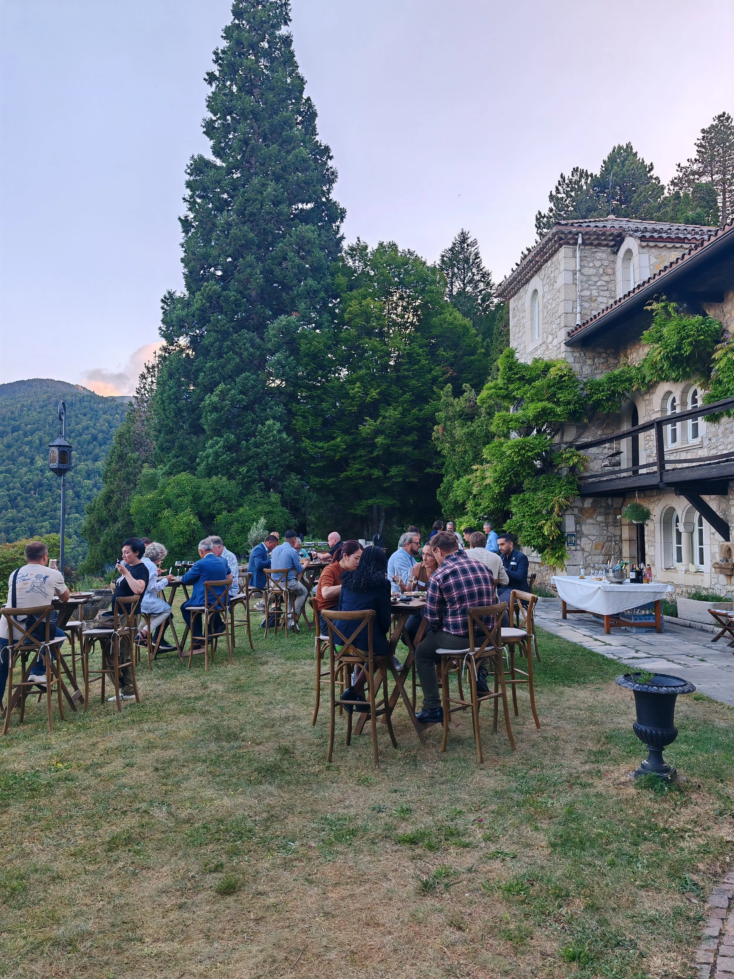 People dining outdoors on a grassy lawn next to a stone building, with trees and mountains in the background.