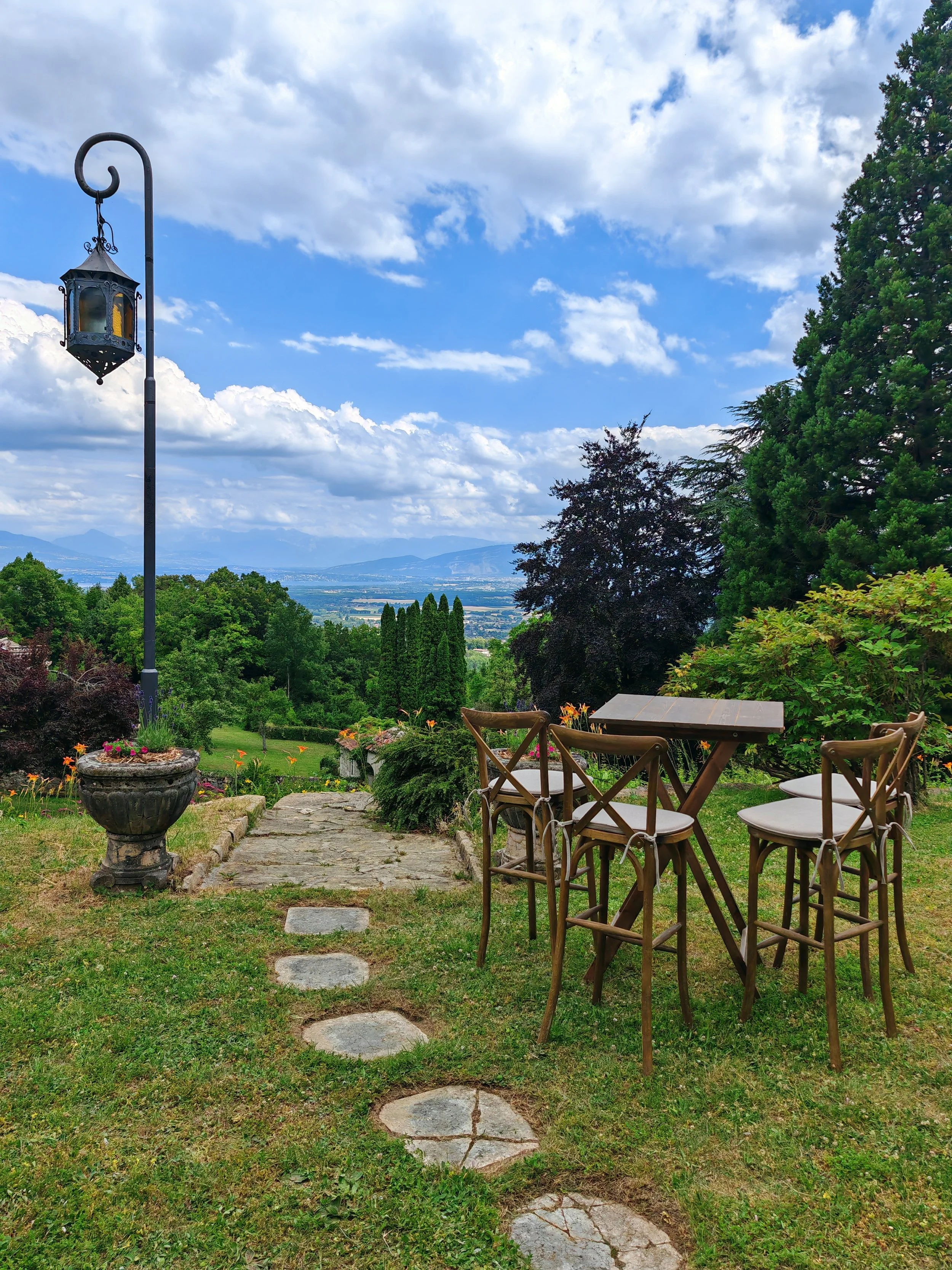 Outdoor garden with stone pathway, wooden chairs and table, lush green trees, potted plants, and a decorative lantern pole with a cloudy sky in the background.