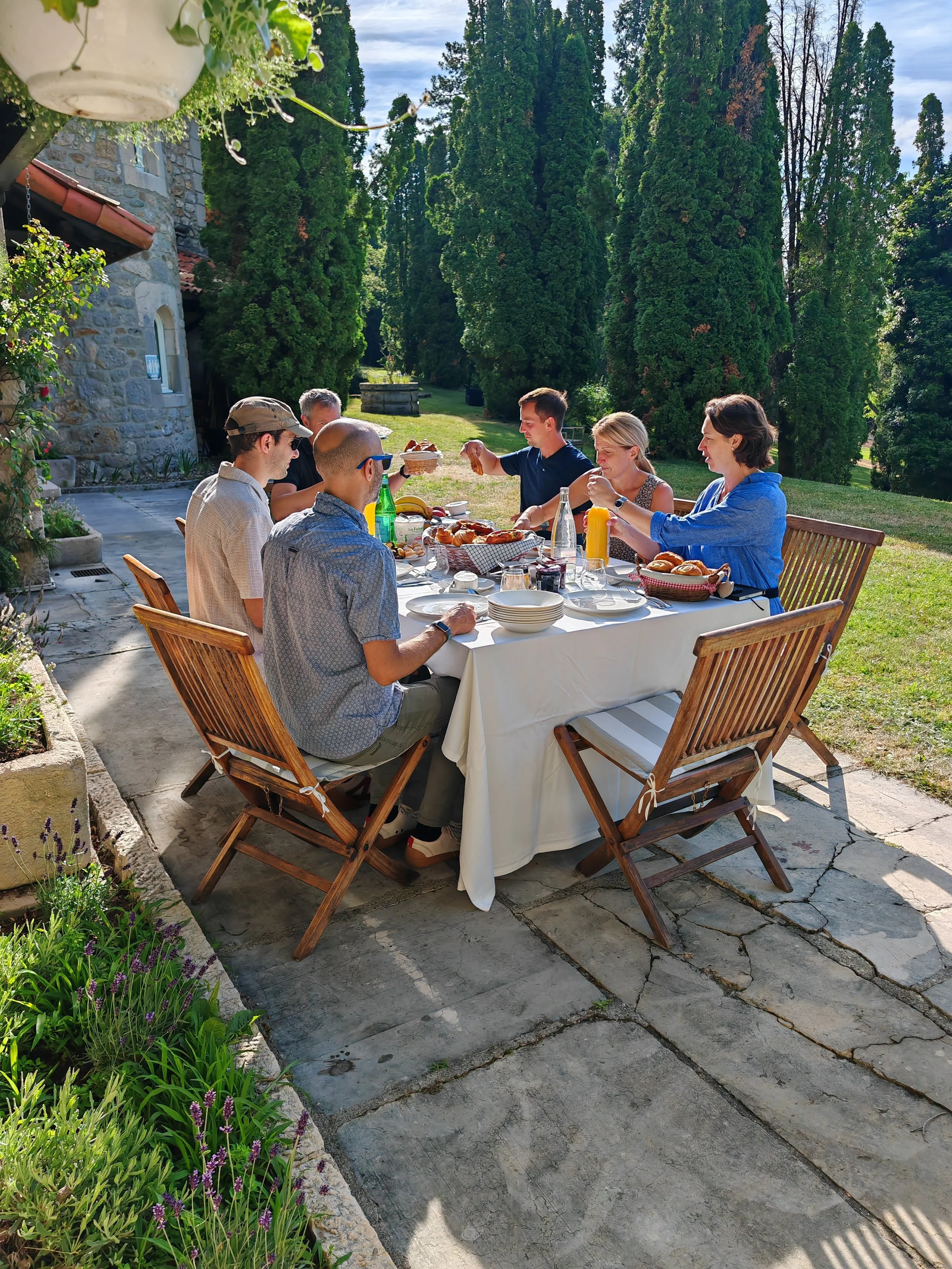 A group of seven people sitting around a table outdoors enjoying breakfast in a garden with tall trees and a stone house in the background on a sunny day.