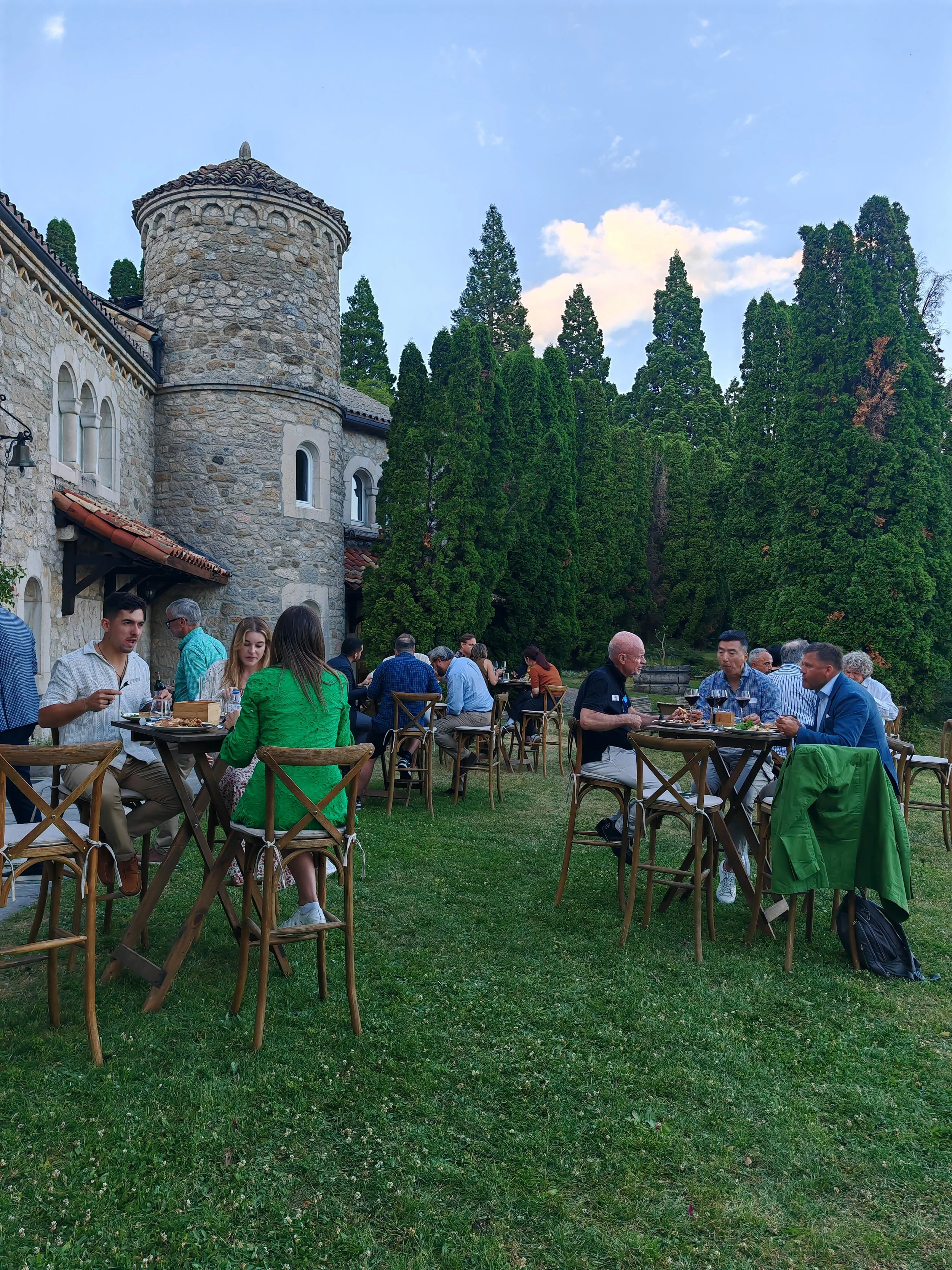 People dining at outdoor tables in front of a stone castle-like building surrounded by tall green trees on a grassy area during daytime.