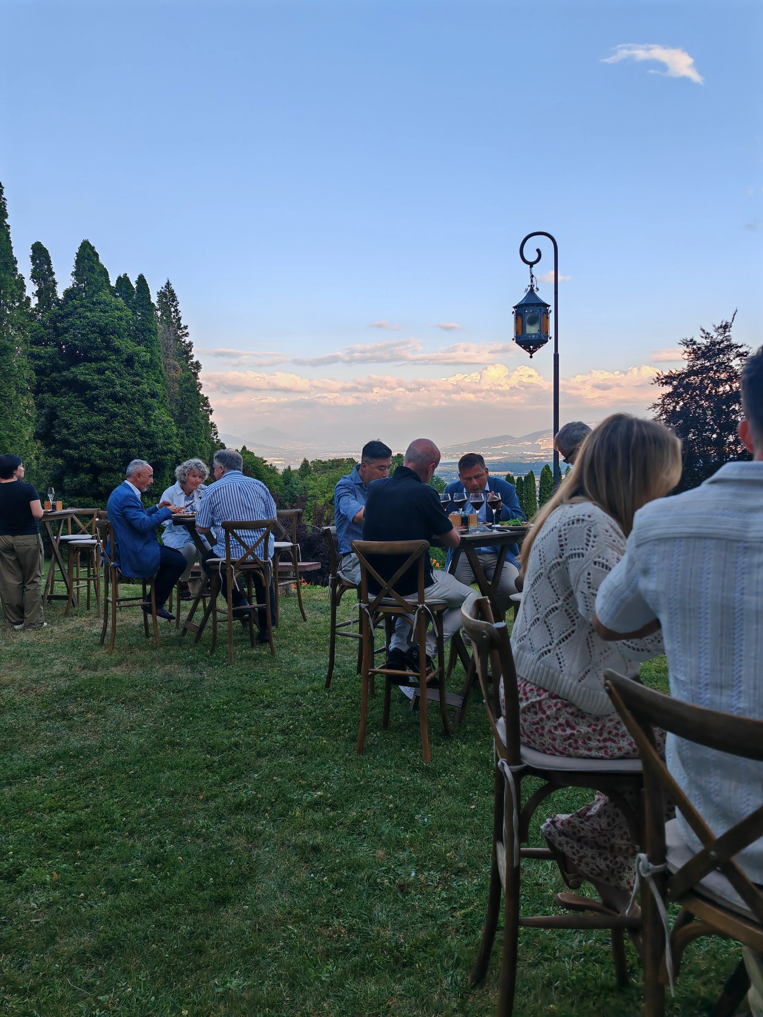 People dining outdoors on a grassy terrace with trees, a sunset sky, and a lantern in the background.