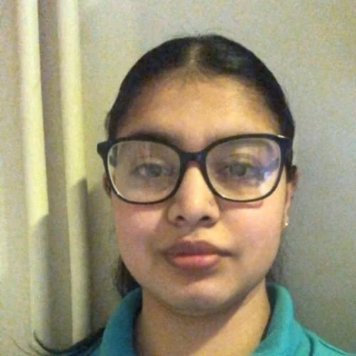 A girl with glasses, dark hair in a ponytail, and earrings, wearing a blue shirt, standing in front of a yellow curtain.