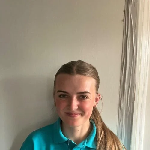 A young woman with long brown hair tied back, wearing a teal polo shirt, standing against a plain wall with a partially visible window and curtains.